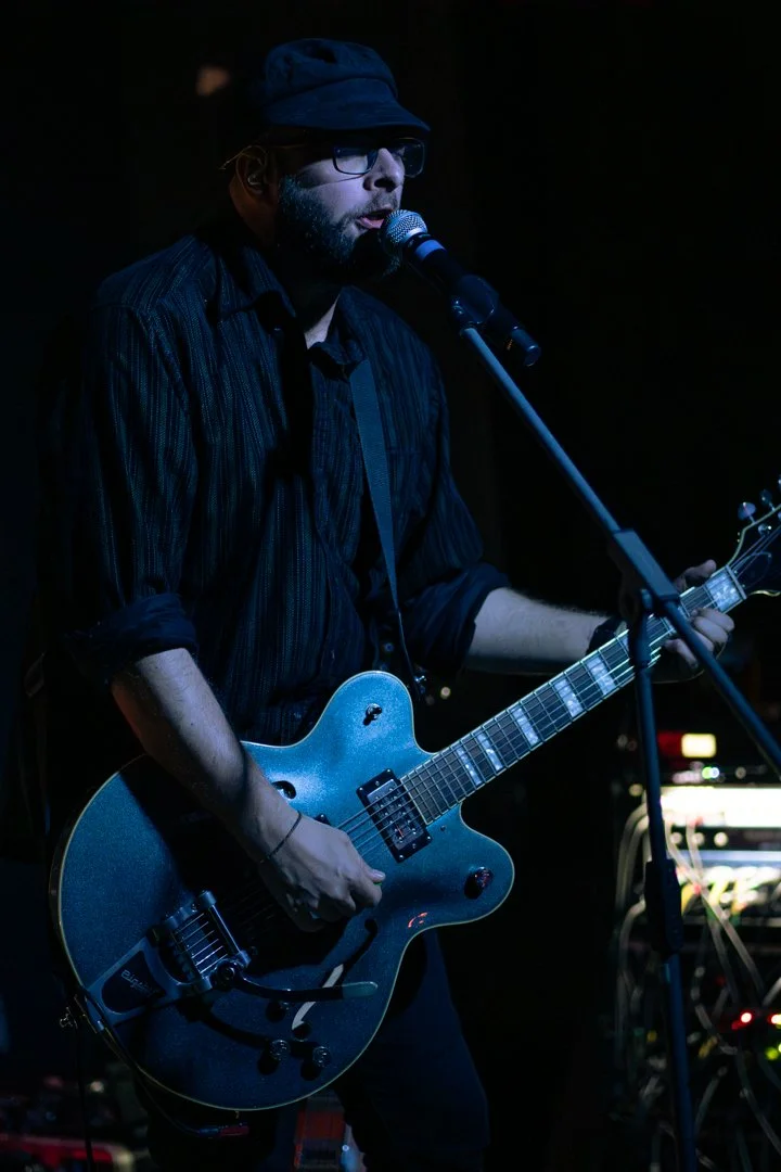 A man wearing glasses, a black hat, and a striped dark shirt is singing into a microphone while playing an electric guitar on stage in a dimly lit setting.