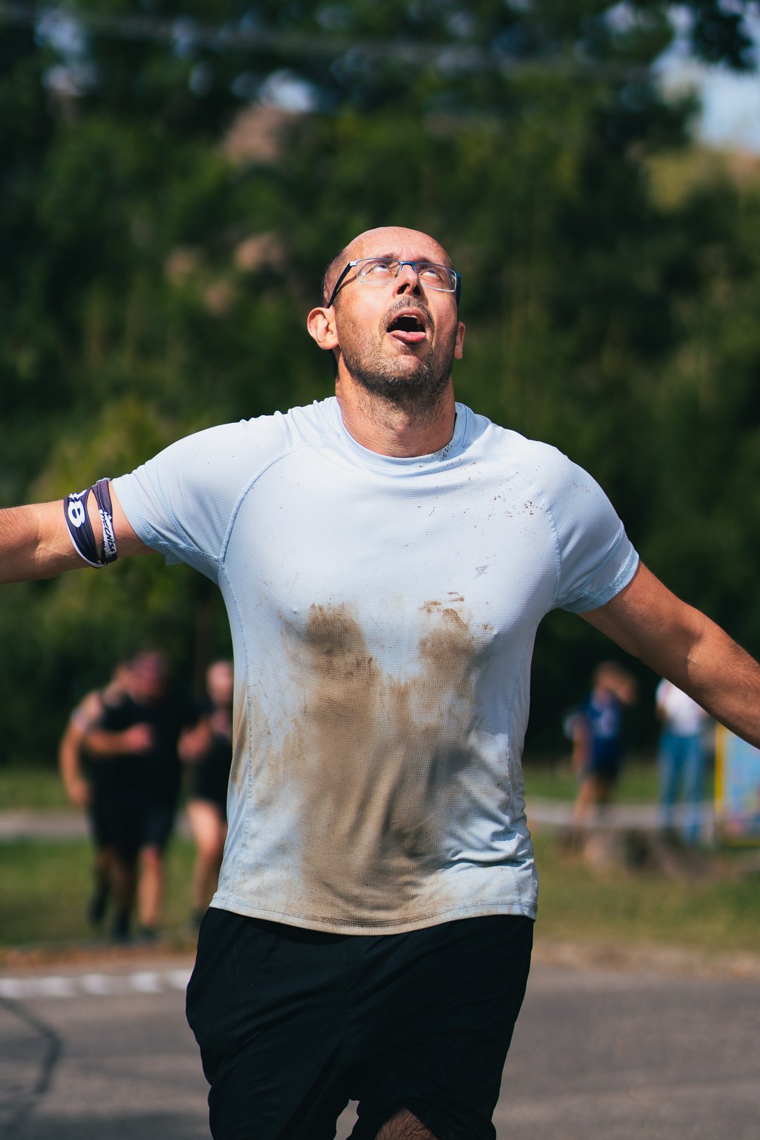 A man with glasses and a beard is running outdoors with his arms outstretched, wearing a dirty white shirt and black shorts, appearing exhausted or triumphant. There are other people and trees in the background.