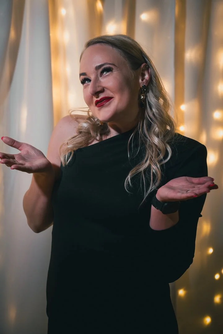 A woman with long, wavy blonde hair, wearing a black one-shoulder dress, smiling and shrugging her shoulders against a backdrop of golden fairy lights.