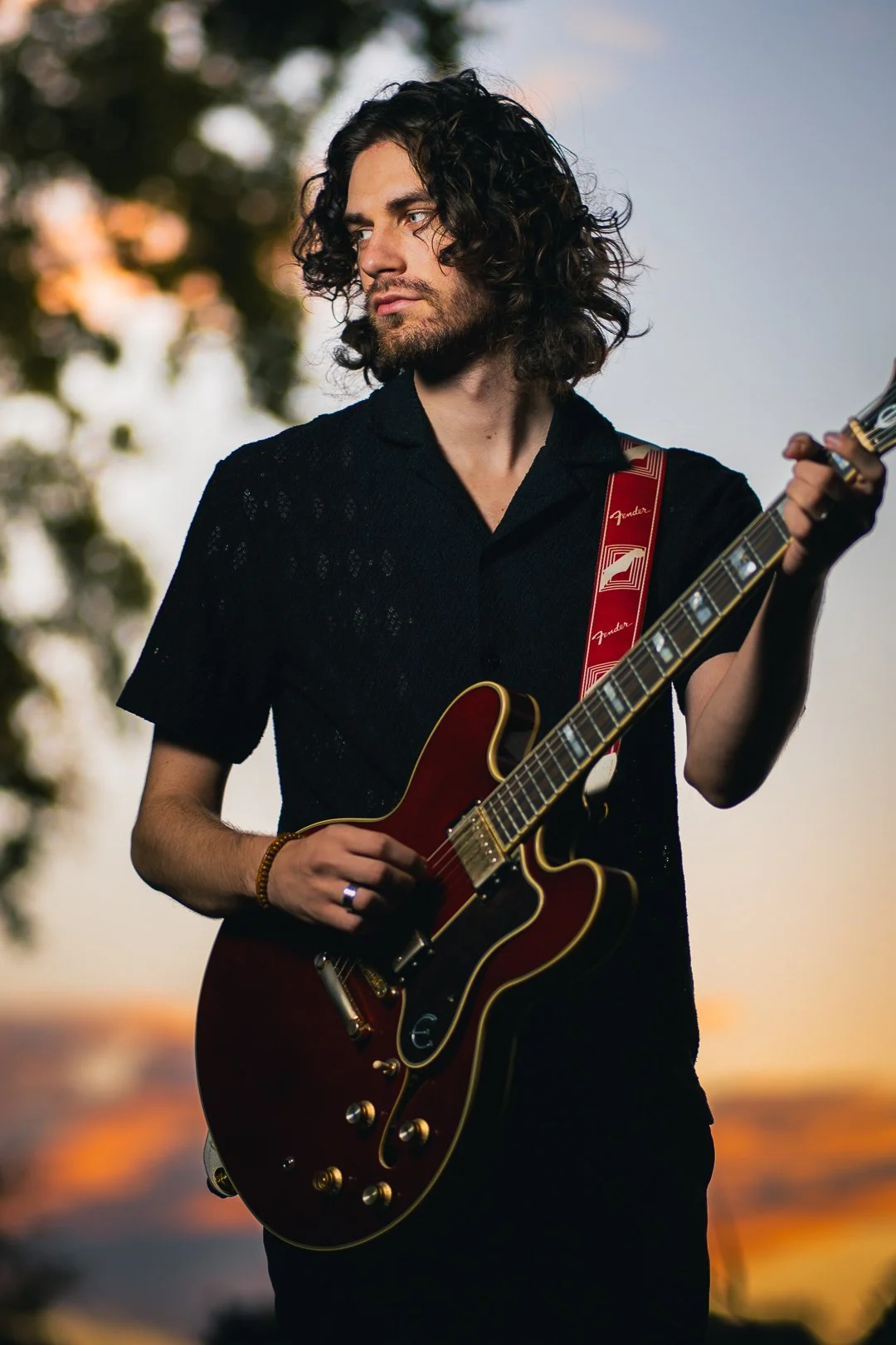 A young man with long curly hair is playing a red electric guitar outdoors during sunset, with a blurred background of trees and a colorful sky.