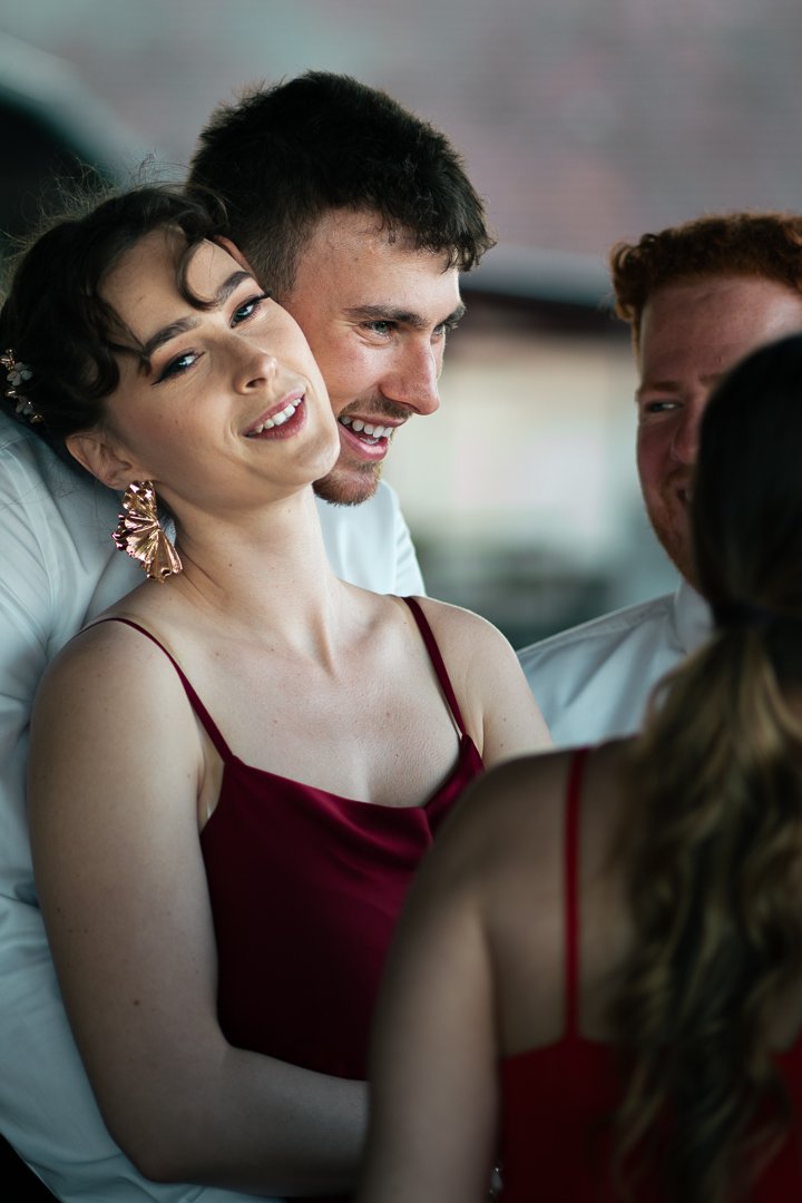 Group of four young adults celebrating, smiling and laughing, with a woman in a red dress in the foreground, at a social gathering or party.