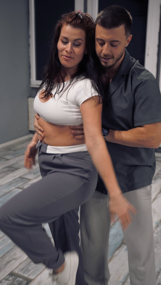 A man and woman dancing closely together in a room with gray walls and a wooden floor.