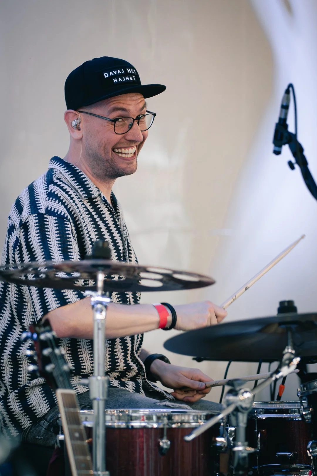 A smiling man wearing glasses, a black cap with text, and a patterned shirt playing drums during a performance.