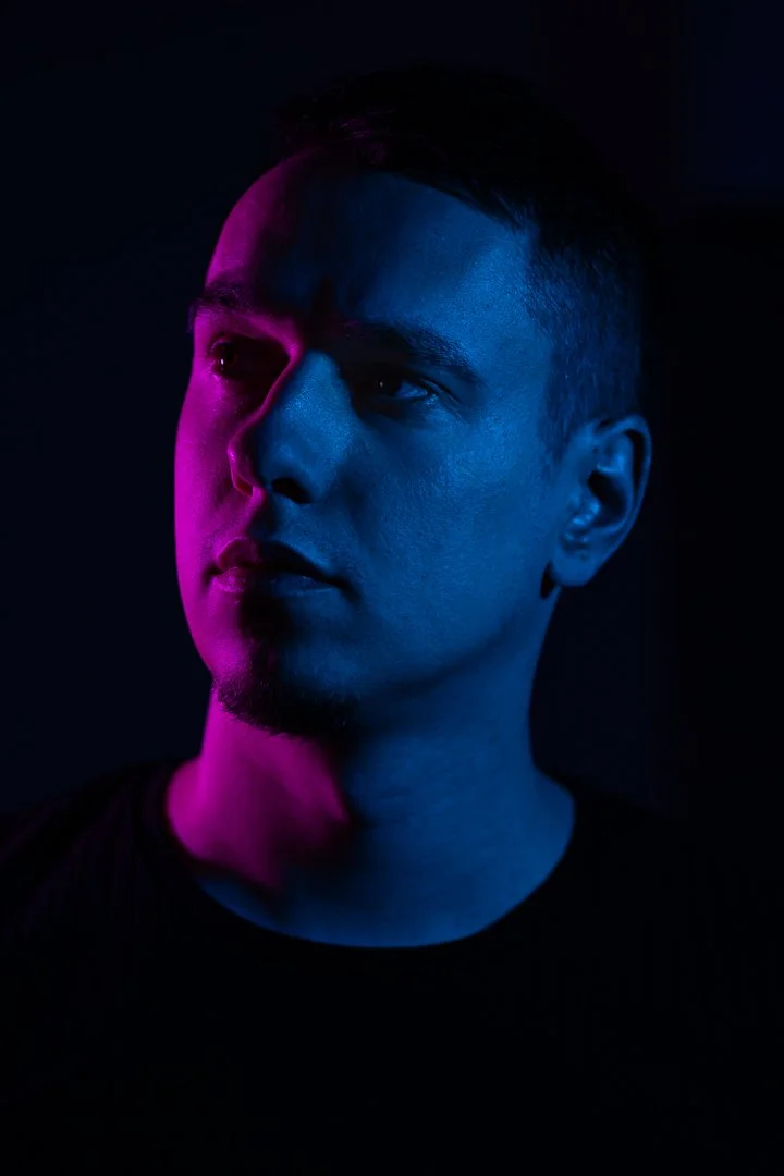 A young man with short dark hair and a goatee looks to the right. The photo is lit with pink and blue lighting, creating a moody, artistic effect.