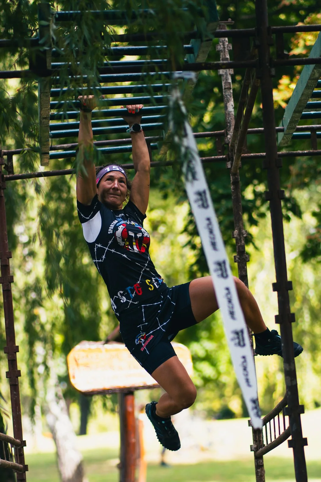 A person participating in an obstacle course, hanging from a bar with one hand, in an outdoor park with green trees.
