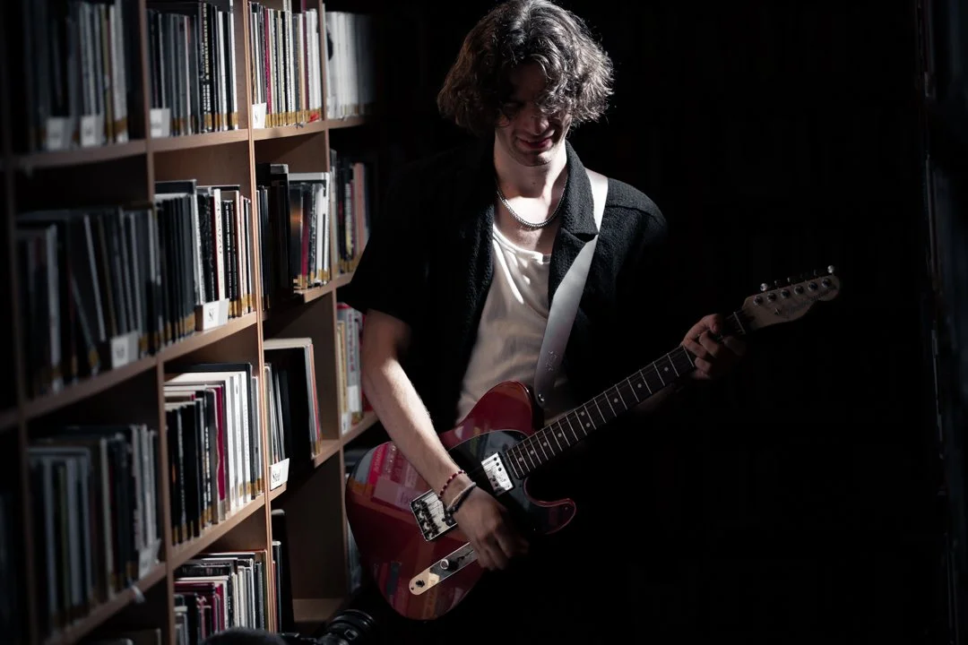 A young person with curly hair playing an electric guitar in a dimly lit library.