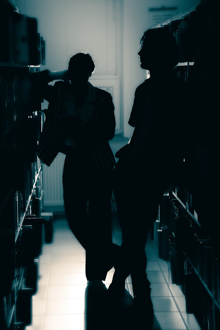 Silhouettes of two people standing in a hallway with lockers, backlit by a window, creating a moody, shadowed atmosphere.