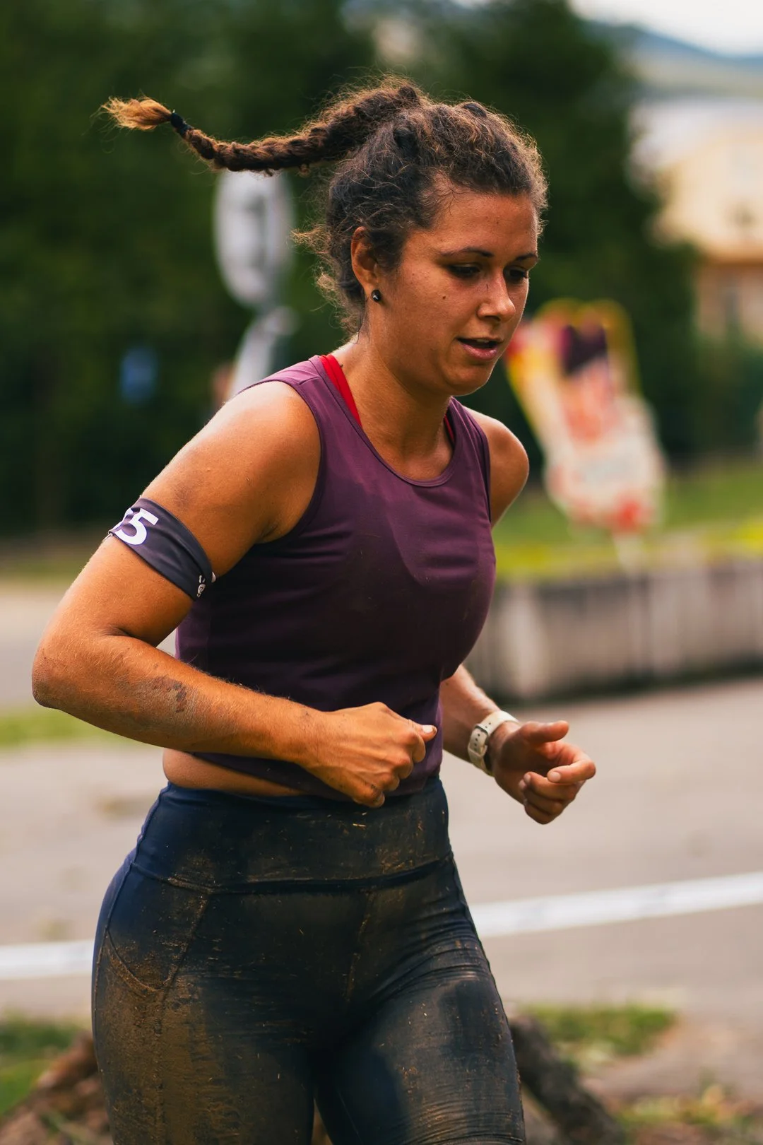 A woman running outdoors with mud on her clothes, wearing a purple top and black leggings, with her hair in a braid.