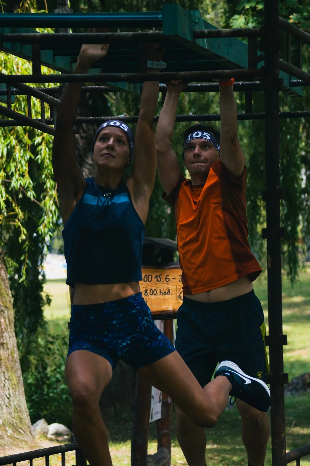 Two runners, a woman and a man, are holding onto a metal structure outdoors, with the woman pulling up while the man does a chin-up. They are surrounded by trees and greenery, and both are wearing headbands and athletic clothing.