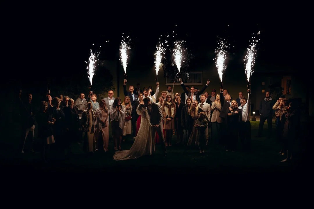 Group of people celebrating with sparklers outside at night, some dressed in formal attire, three people in the center are kissing, likely at a wedding reception.