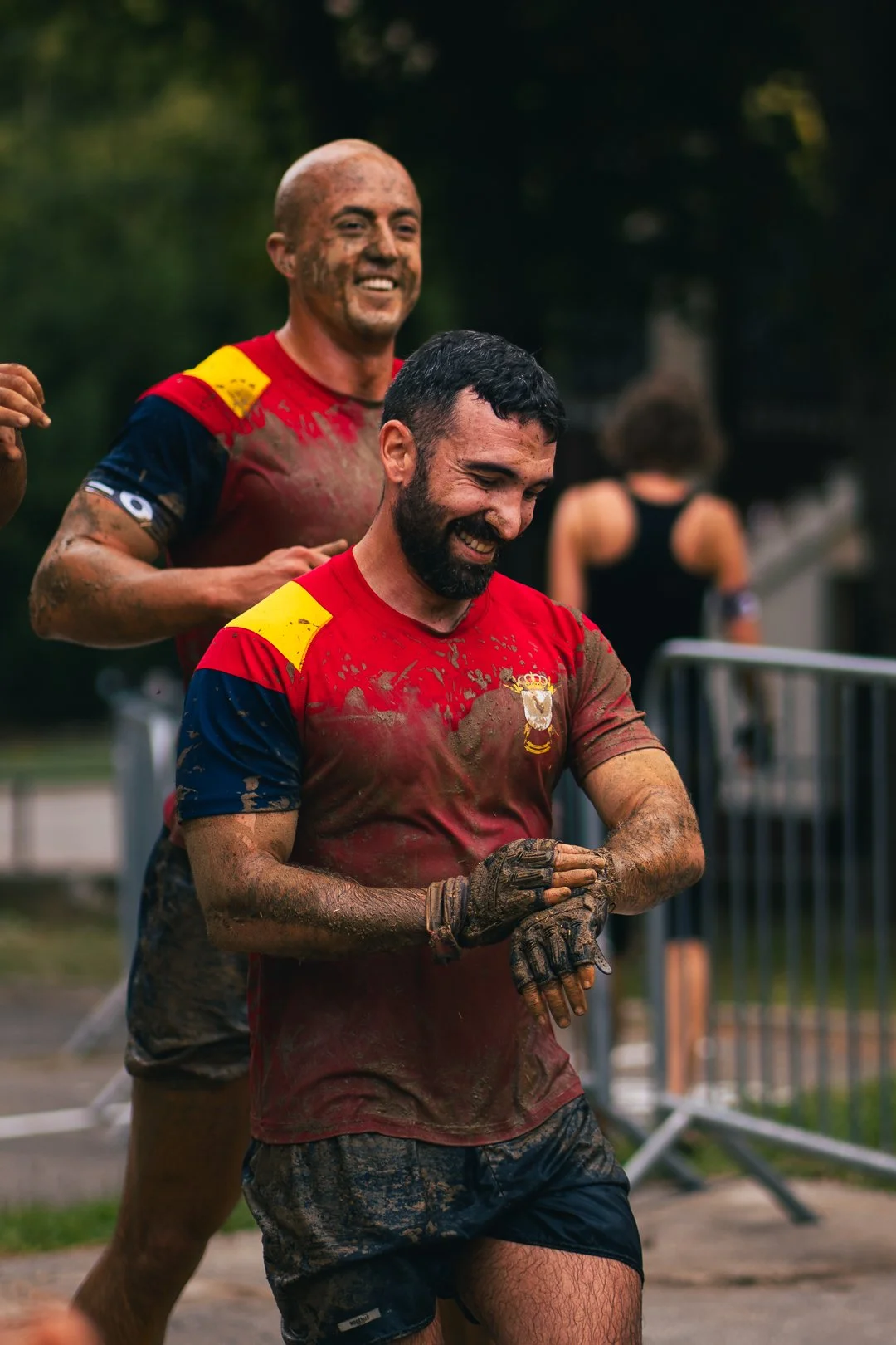 Two smiling men with muddy sports uniforms running outdoors during a muddy race, with a metal barrier in the background.