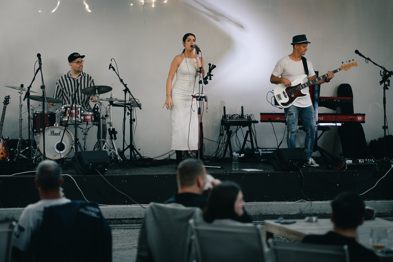 A band performing live on stage with three musicians: a drummer on the left, a female singer in the center, and a male bassist on the right. Audience members are visible in the foreground.