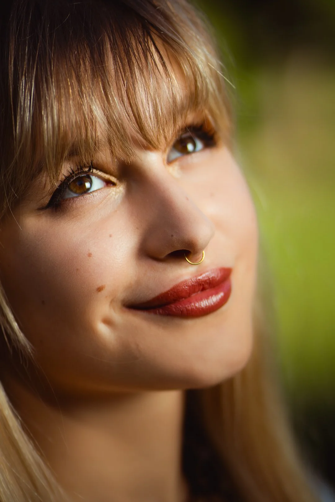 Close-up of a woman with light brown hair, brown eyes, a septum piercing, and red lipstick, smiling outdoors with a blurred green background.