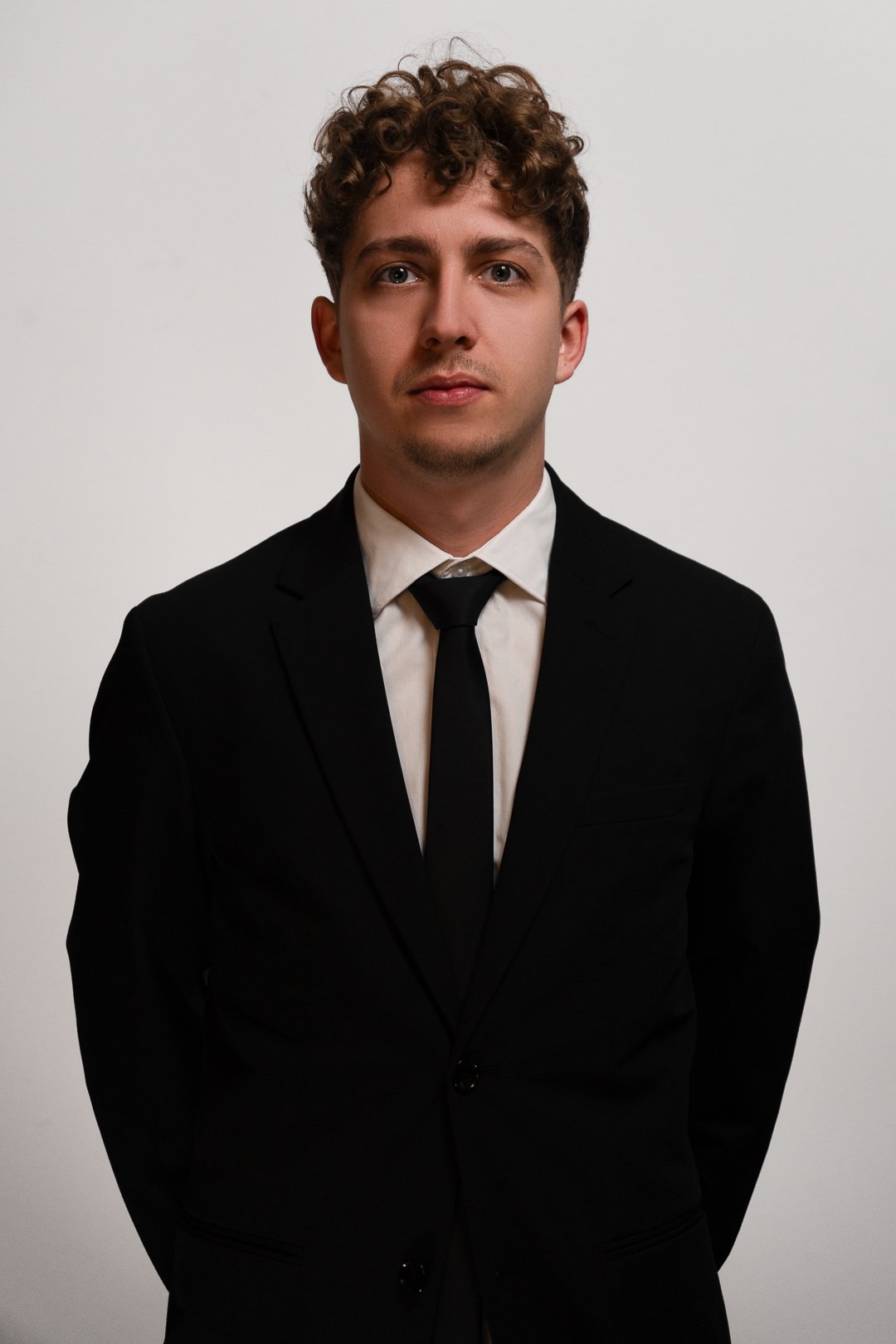 Young man with curly hair wearing a black suit, white shirt, and black tie standing against a plain white background.