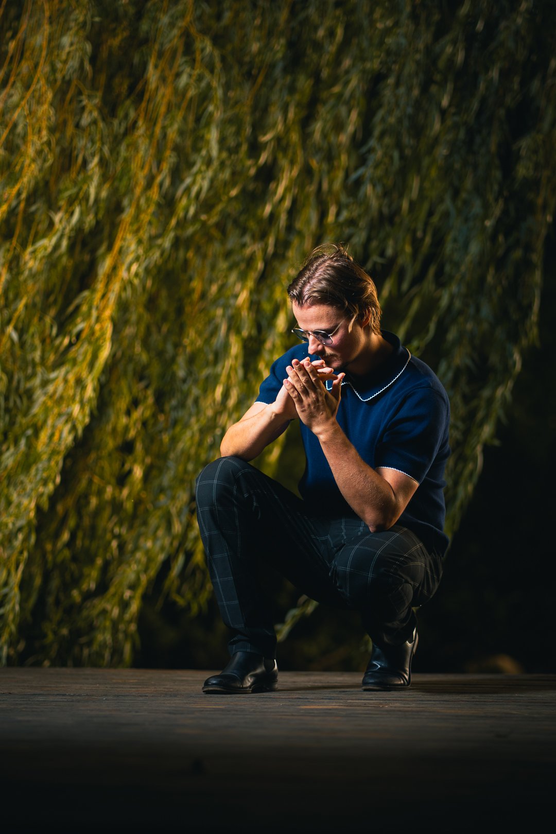 A man crouching on a dark wooden platform at night, wearing glasses, a blue shirt with a white collar, and checkered pants, with tall green plants in the background.