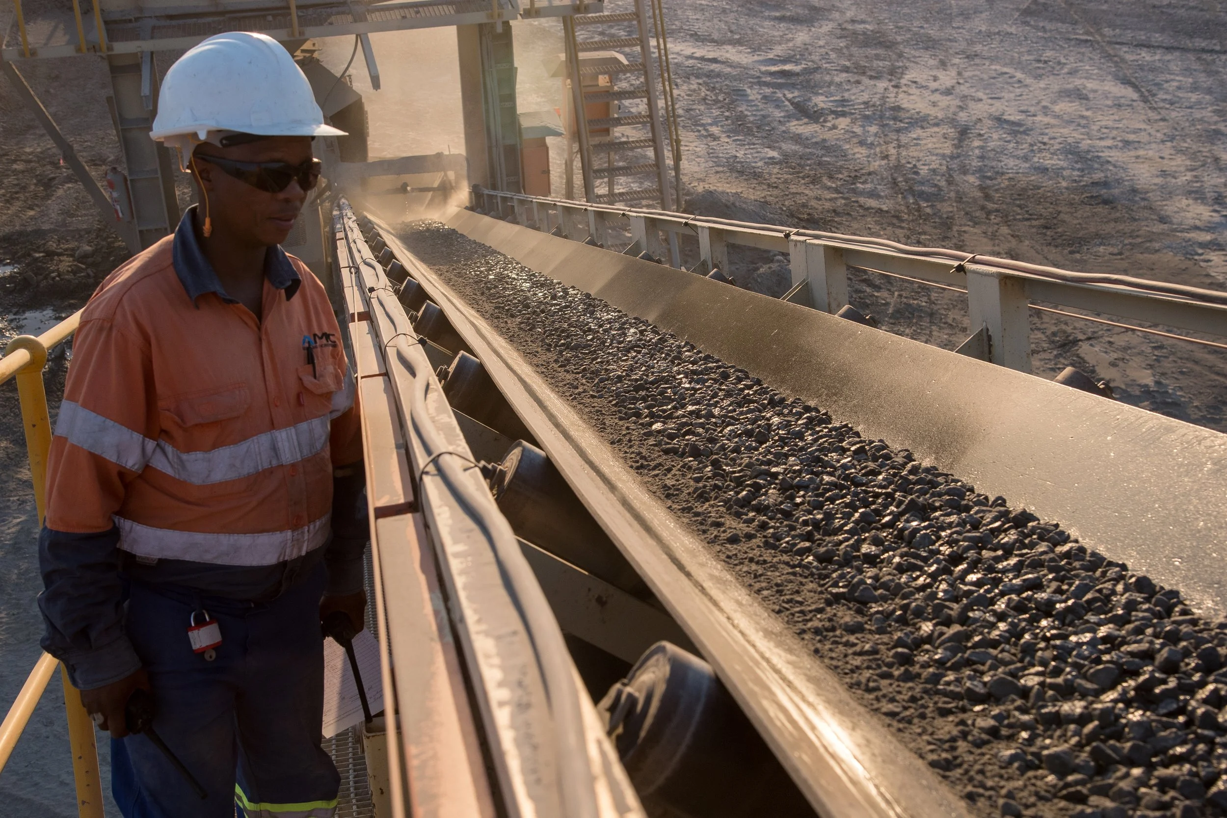A worker wearing safety gear, including a white hard hat and sunglasses, inspecting an industrial conveyor belt carrying a load of coal or similar material.