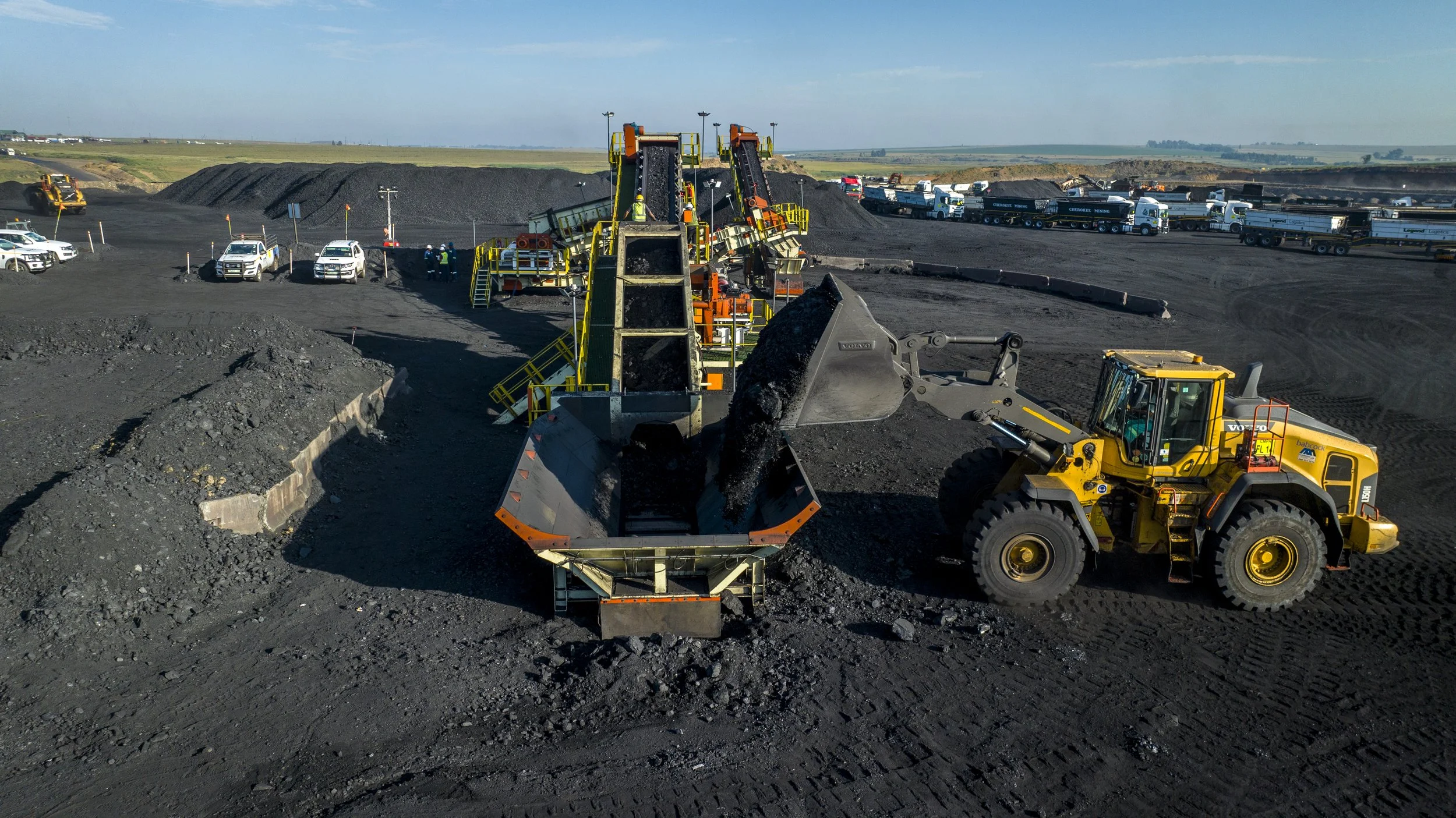 Construction site where a large yellow front loader is loading black asphalt into a paving machine, with piles of asphalt and multiple construction vehicles in the background under a clear sky.
