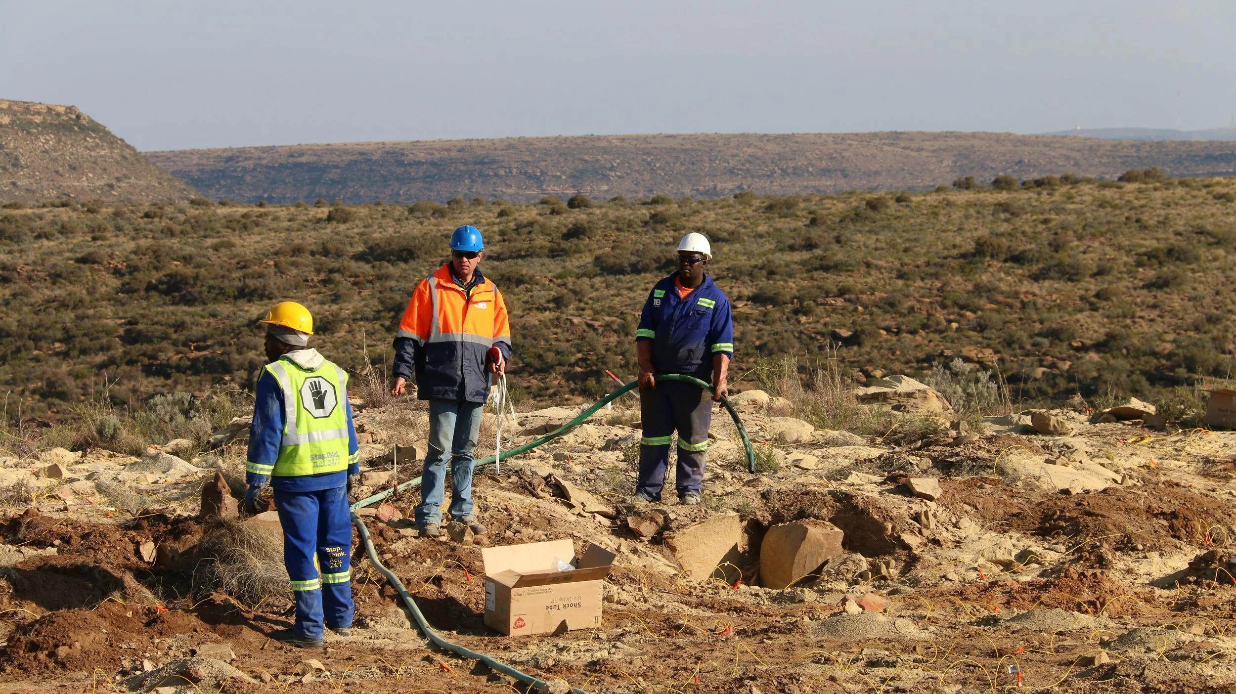 Three workers in safety gear, including helmets and reflective vests, working on a construction site in a desert landscape with rocky ground and distant hills.