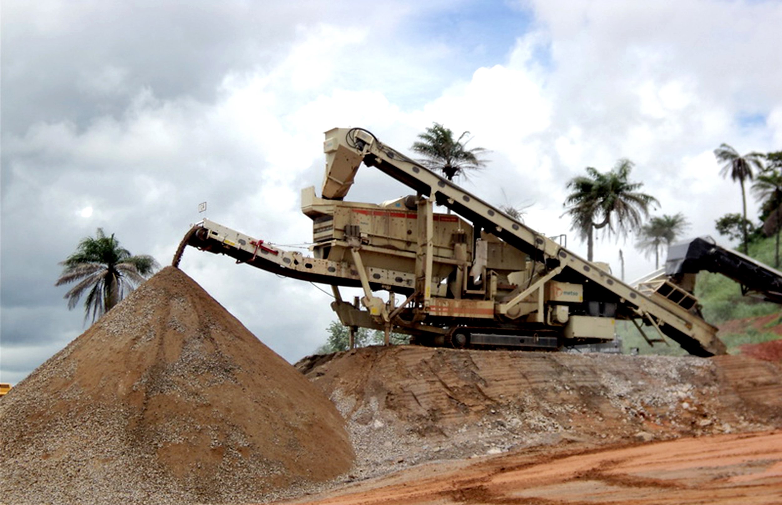 A bulldozer moving dirt on a construction site with palm trees and a cloudy sky in the background.