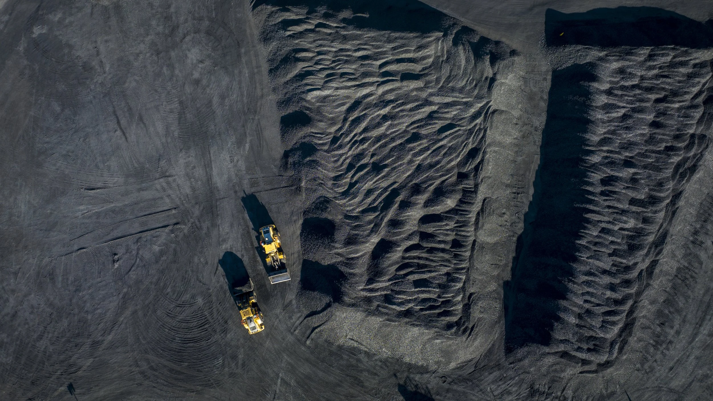 Three yellow construction dump trucks on a dark, textured dirt or gravel surface with tire tracks and small mounds of dirt.