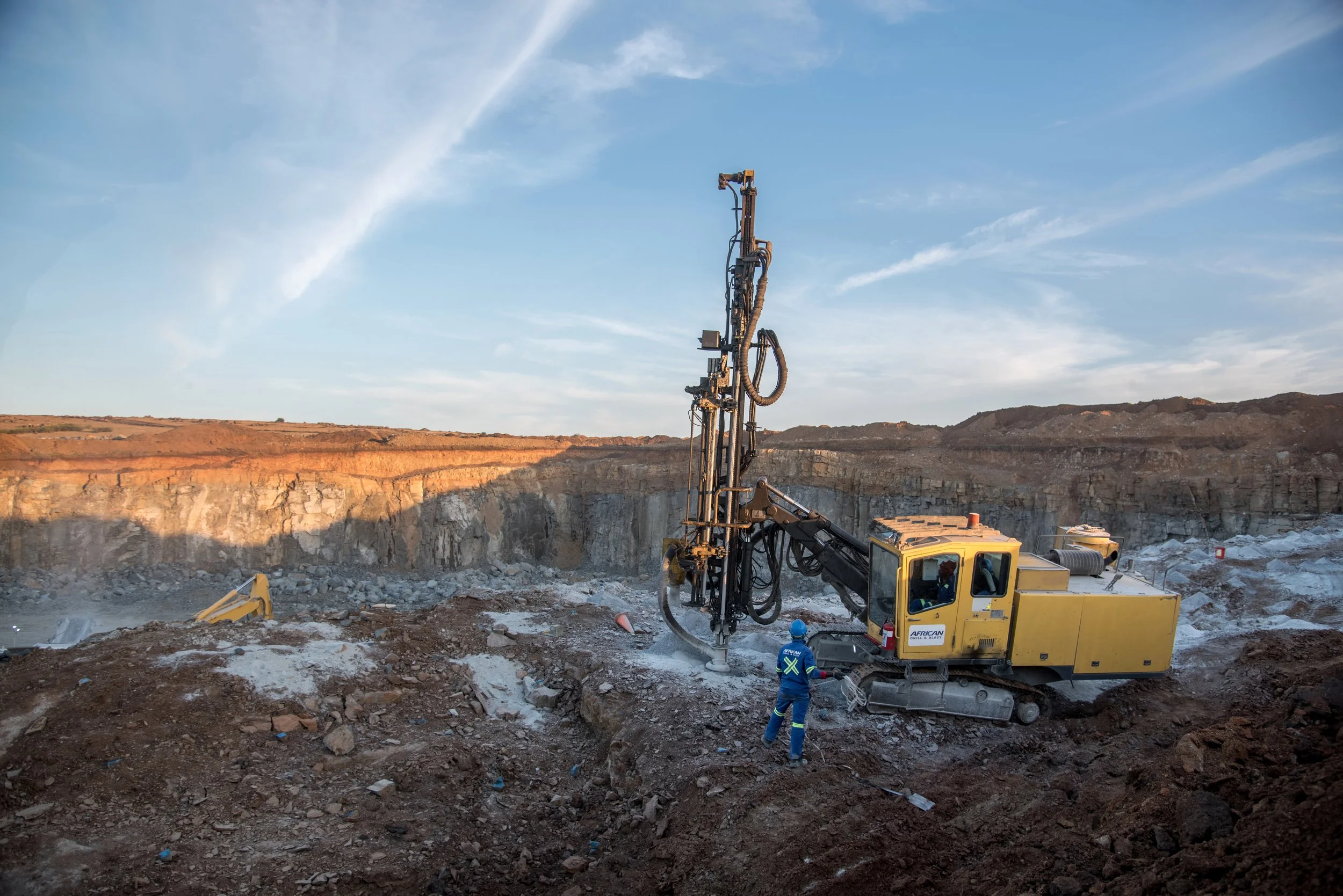 A yellow drilling machine operating at a mining site with a worker standing nearby, geological formations in the background, and a clear sky.