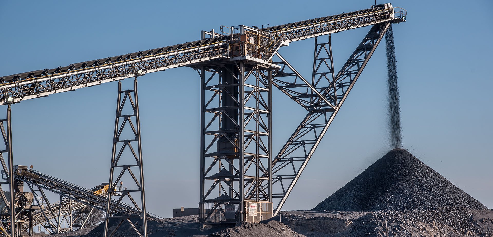Industrial gravel or coal conveyor system transporting material into a large pile below under a clear blue sky.