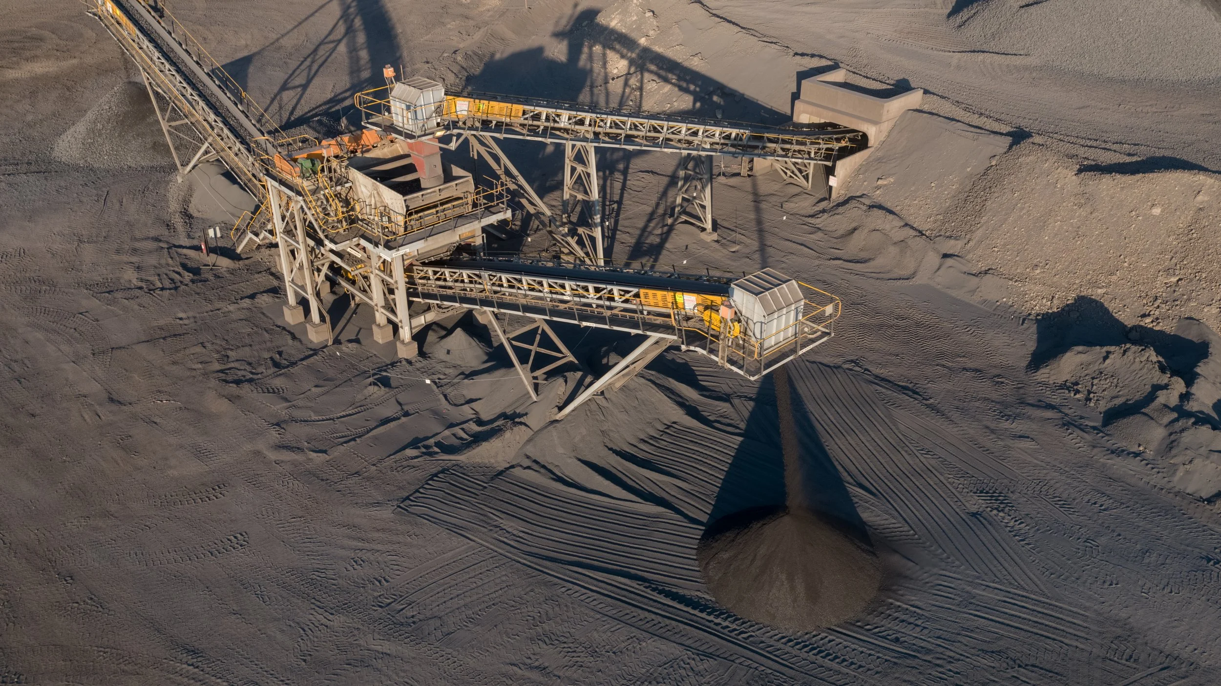 An industrial conveyor system processing and transferring loose material, likely gravel or crushed stone, into a pile on the ground at a quarry or mining site.