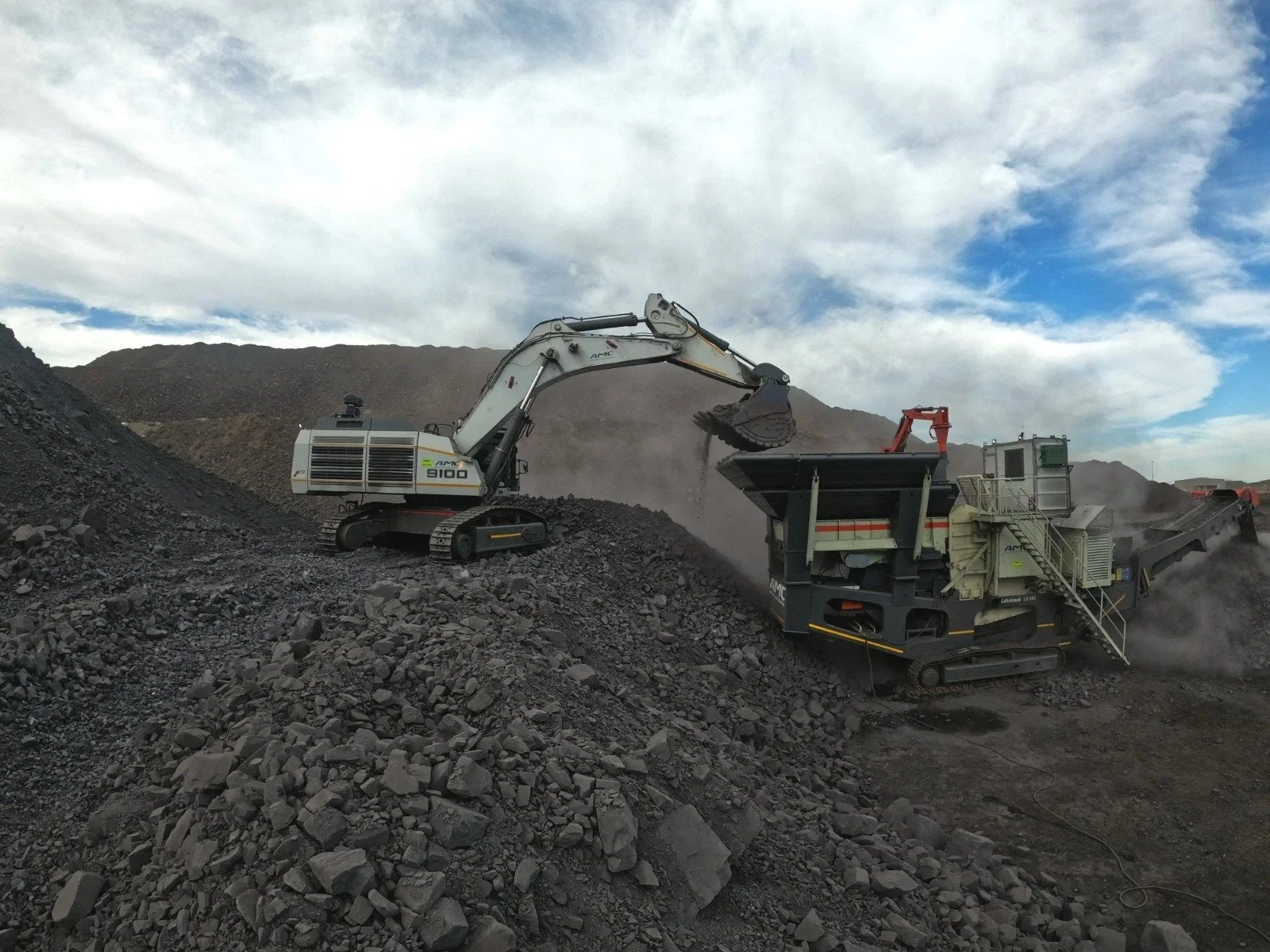 A construction excavator dumping a load of gravel into a mobile crushing machine while working on a mountainous landscape with a cloudy sky.