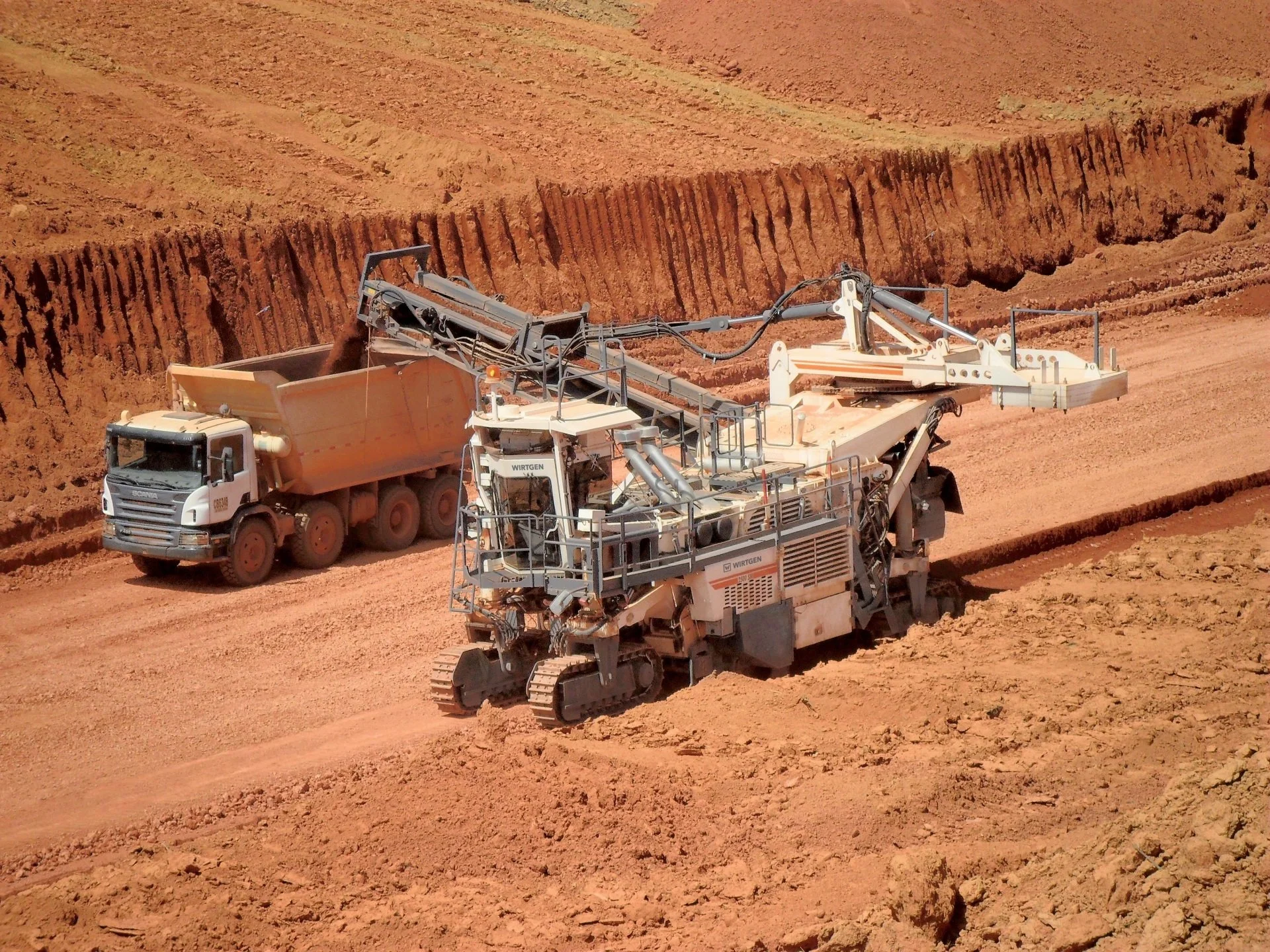 Heavy machinery working on a dry, dusty red earth construction site, with a large tracked excavator and a dump truck.