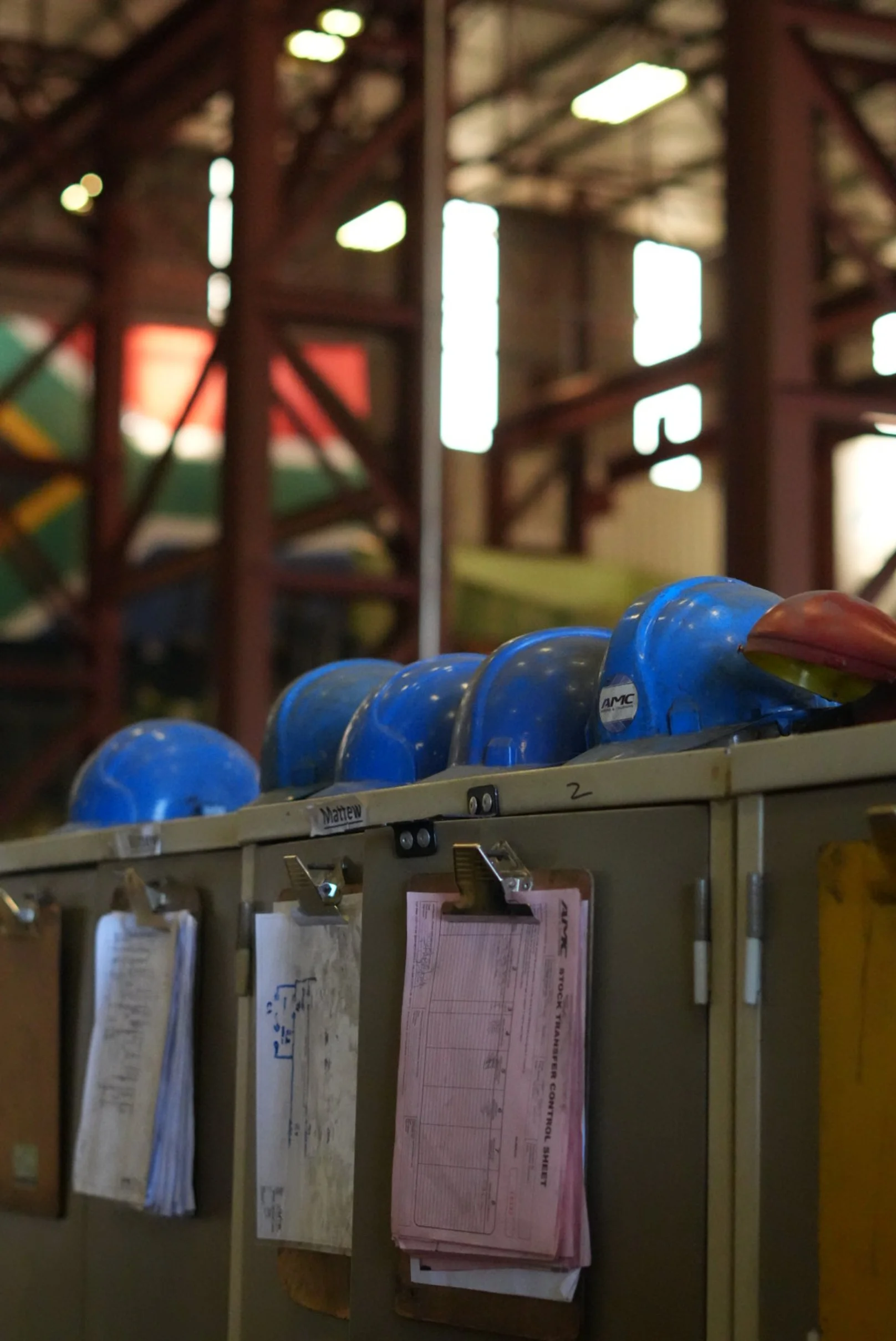 Several blue safety helmets lined up on a locker in an industrial setting with metal structures and windows in the background.