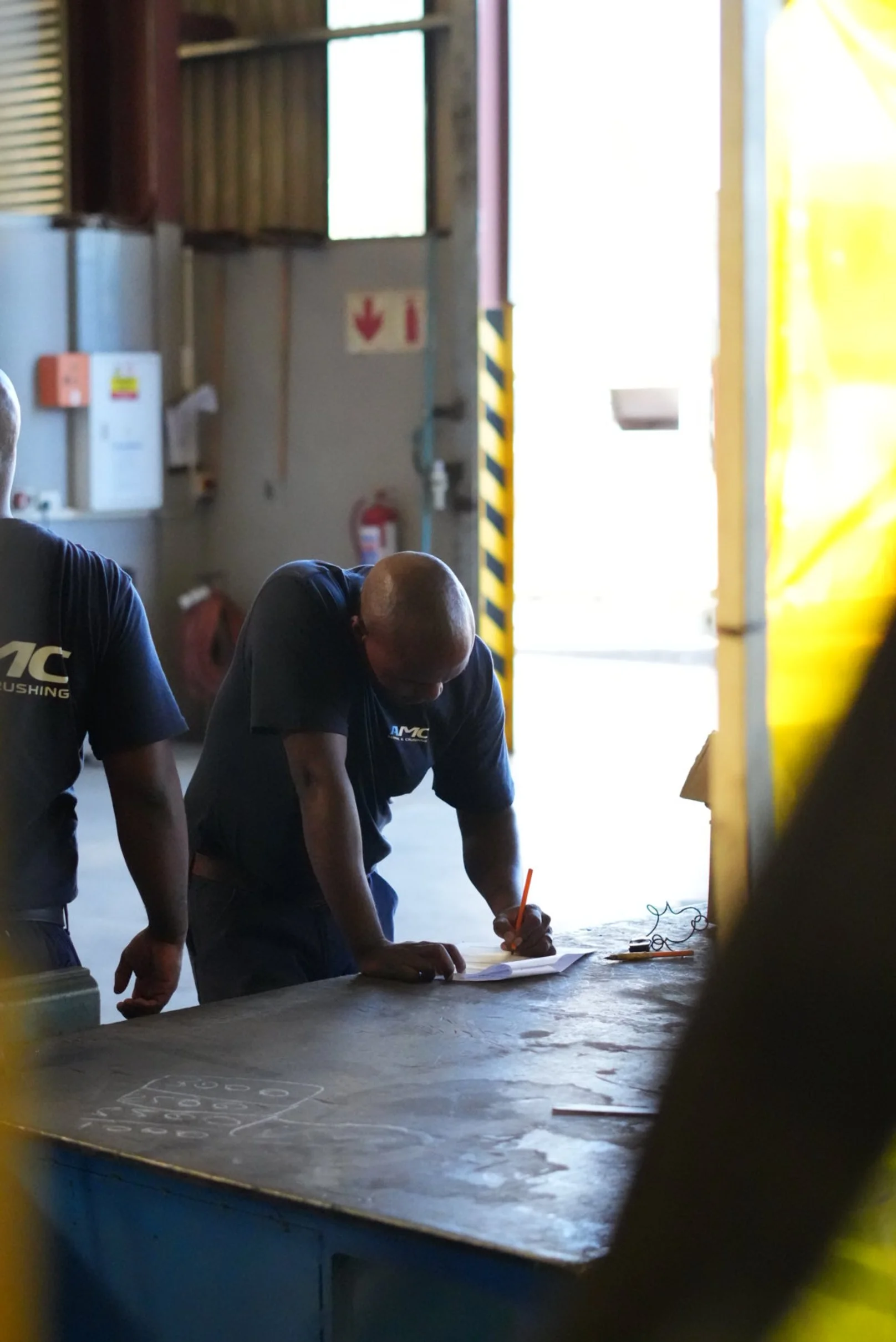 Man leaning over a table, writing on a piece of paper with a pencil. Another man standing beside him. Indoor setting with industrial equipment and bright light coming from the background.