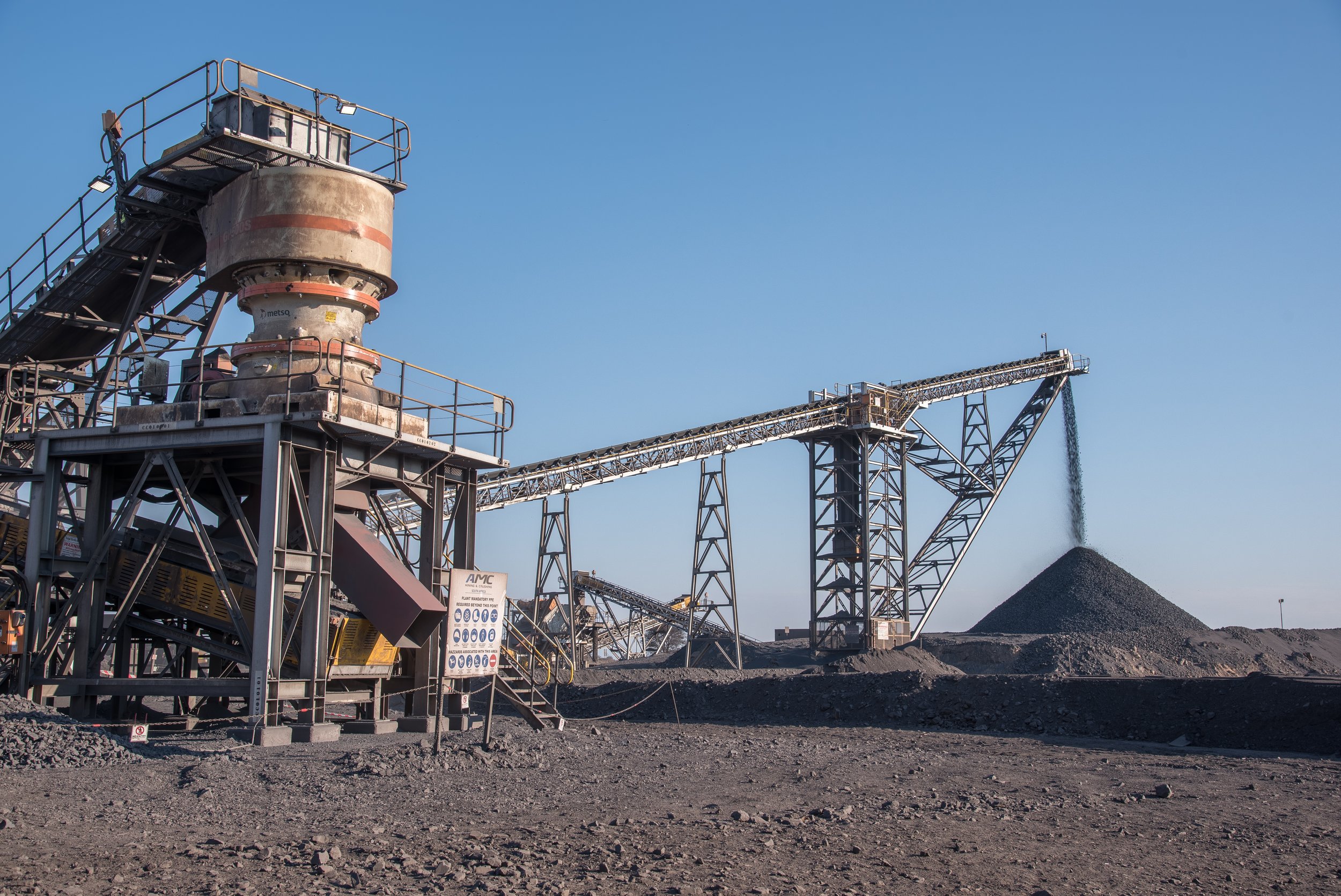 Open-pit mining equipment with conveyor belts and piles of extracted mineral or ore against a clear blue sky.