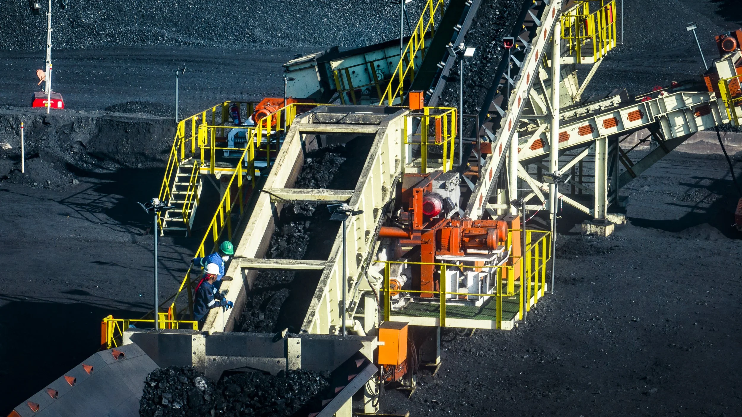 Mining operation with a conveyor belt transporting coal, workers in safety helmets and vests inspecting the machinery.