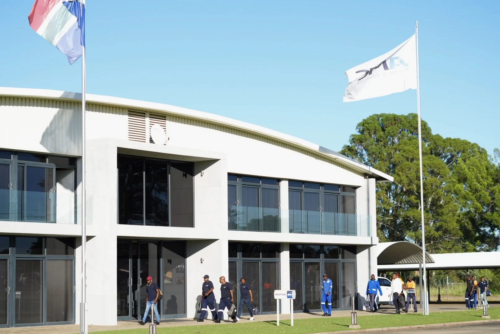 People walking outside a modern building with glass windows and flags flying on poles, one flag with a logo and the other with orange, white, and blue colors, against a background of trees and a clear blue sky.