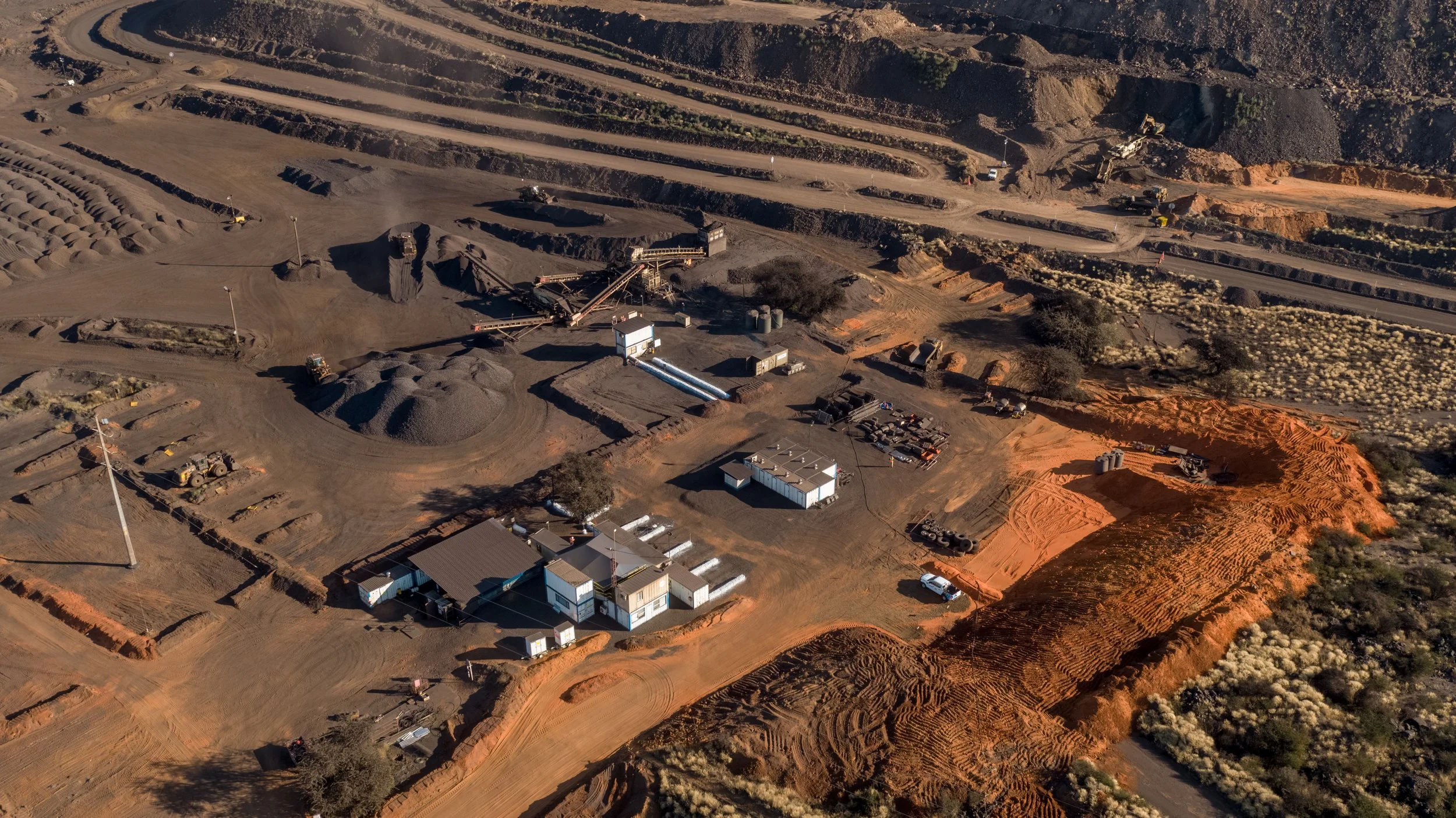 Aerial view of an industrial mining site with conveyor belts, piles of extracted material, construction equipment, and terraced terrain with orange soil and surrounding desert vegetation.