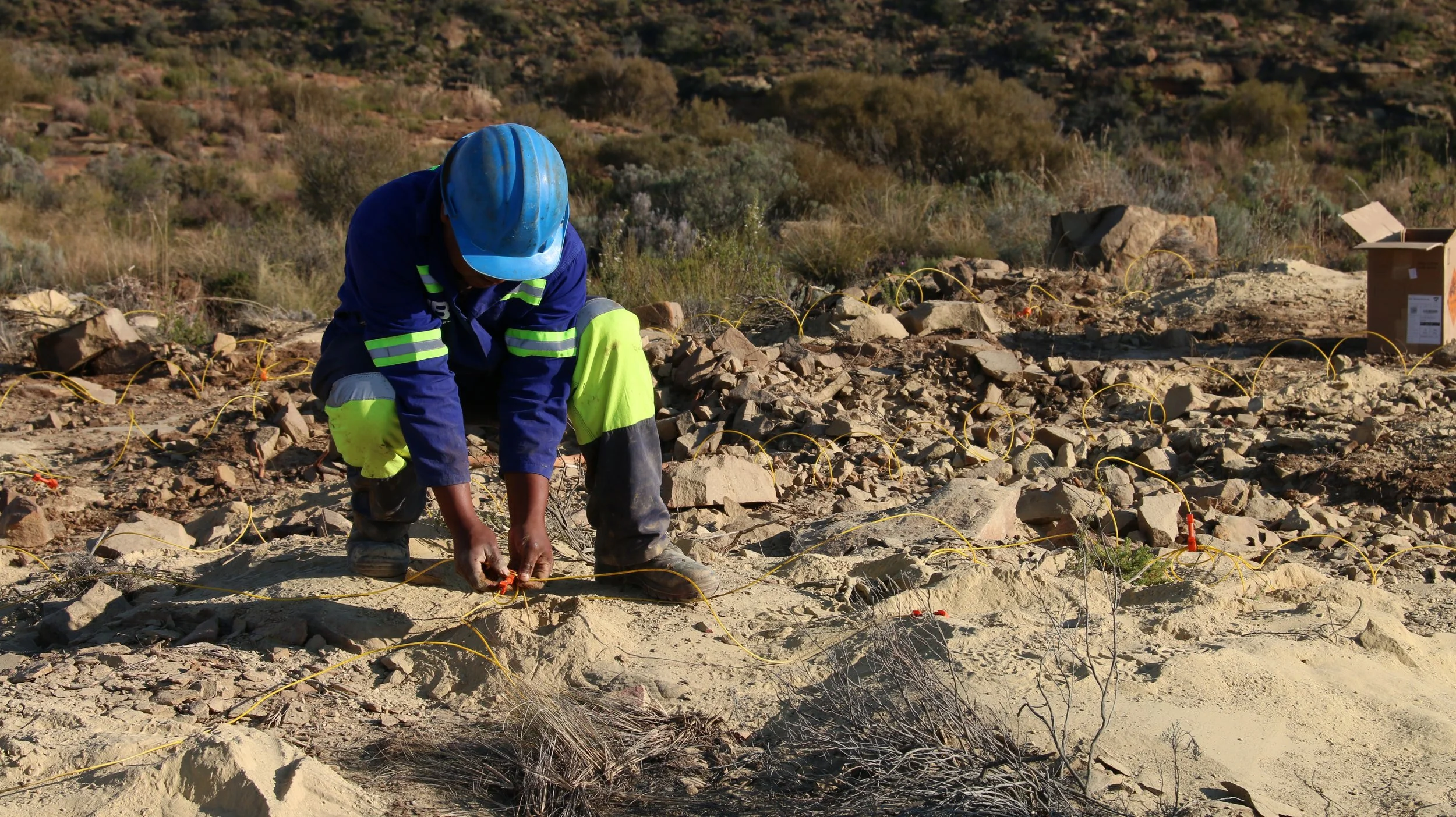 A worker in a blue safety helmet and high-visibility clothing is working on a rocky, desert landscape, installing or inspecting underground cables or wiring with yellow protective wires