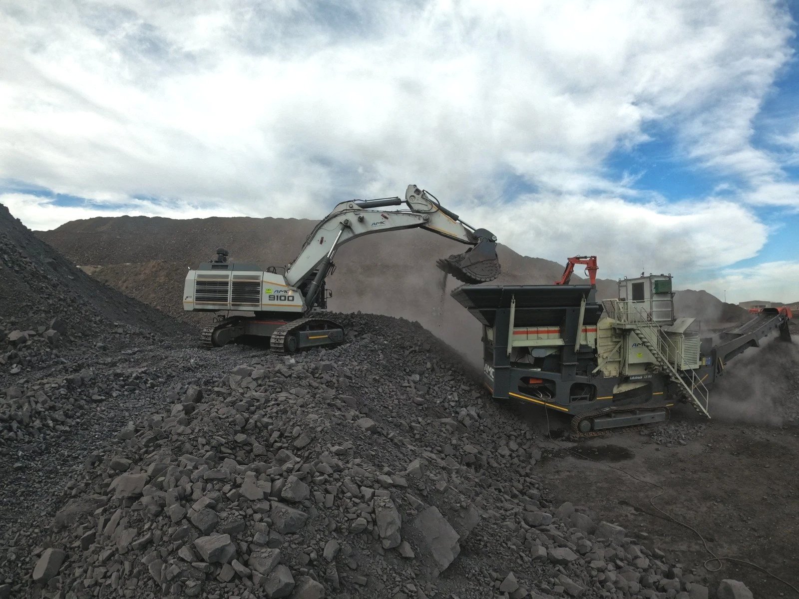Excavator loading rocks into a mobile crusher on a rocky quarry, with cloudy sky and mountains in the background.