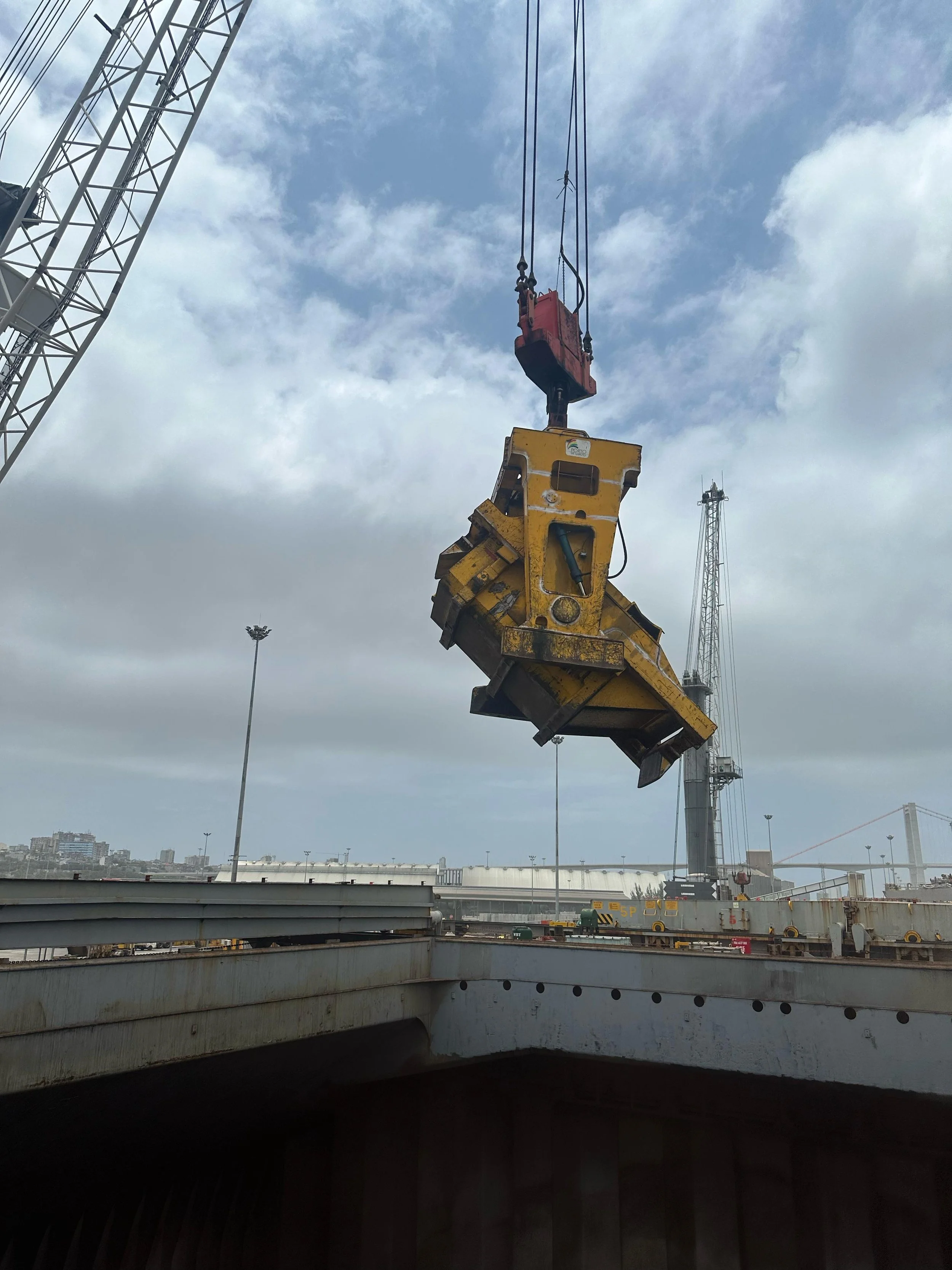 A large construction crane lifting a heavy yellow mechanical component in a construction yard with cloudy sky and distant cityscape in the background.