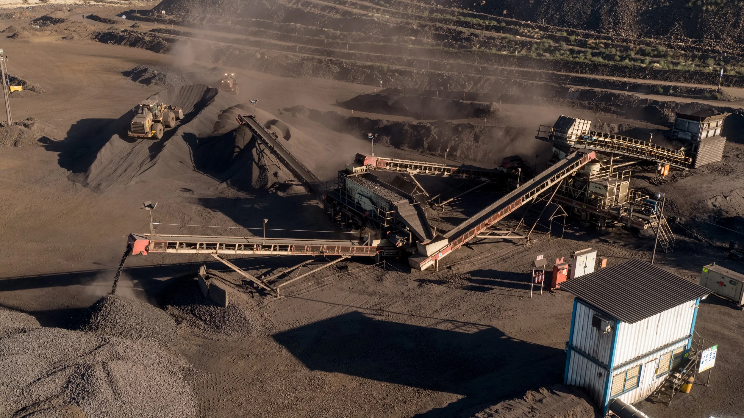 Aerial view of an active gravel or mineral processing plant with crushing and conveyor equipment, several piles of gravel, and bulldozers working amidst dust clouds in a rugged industrial landscape.