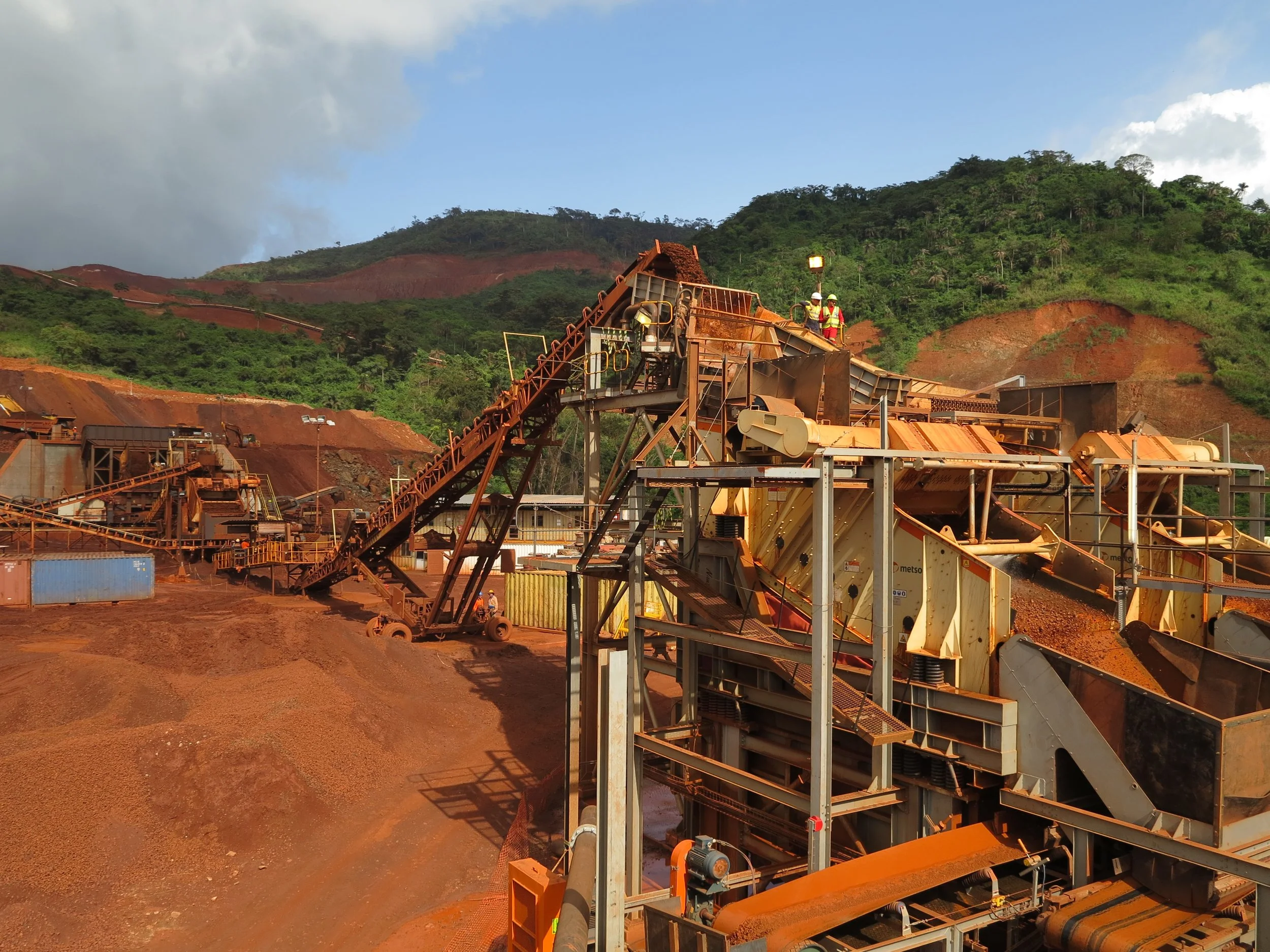 Large industrial mining equipment working in a red dirt quarry with green hills in the background, and workers with safety gear on top of the machinery.