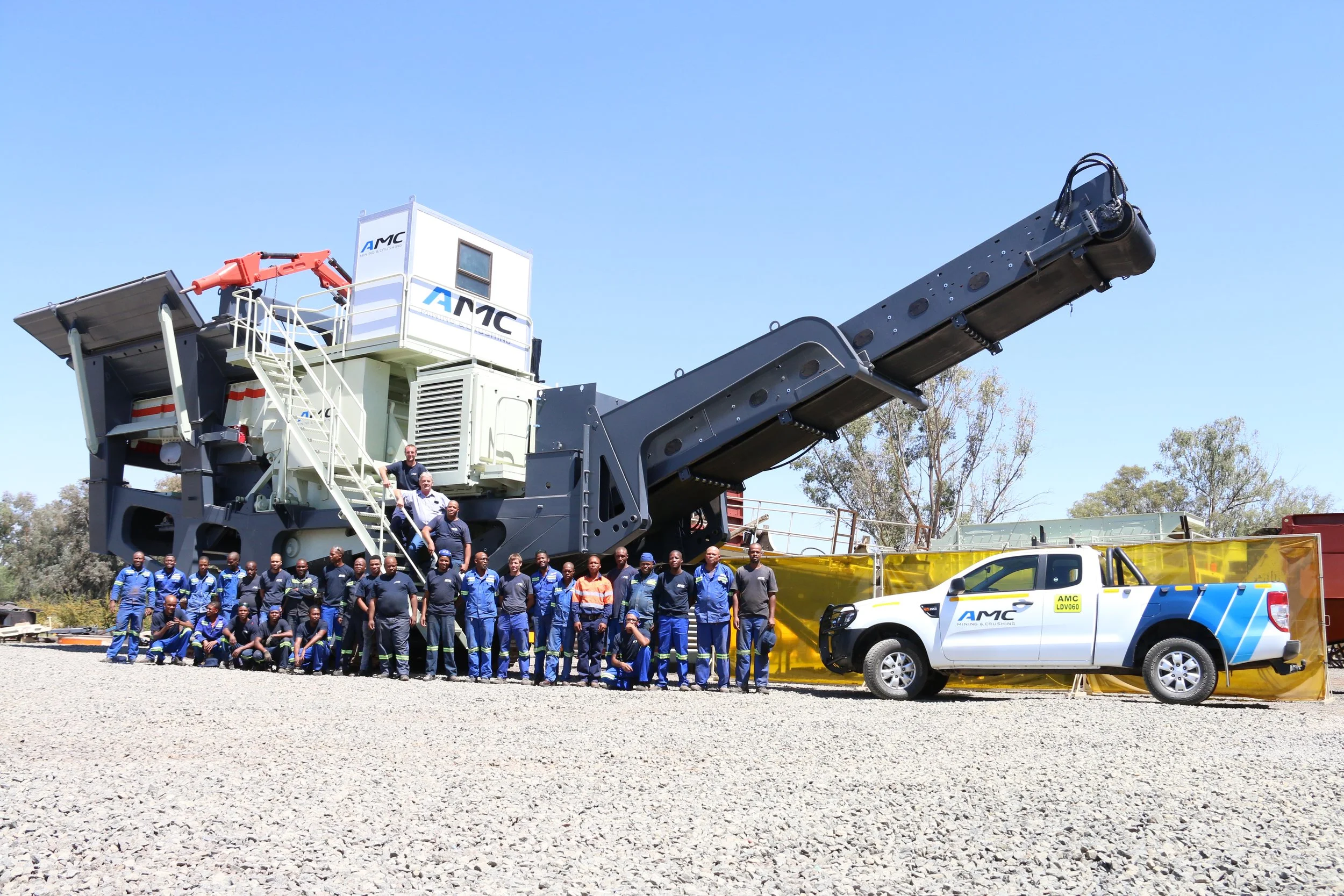 Group of workers in uniforms standing in front of large mining crusher machine and pickup truck with AMC logo, outdoors on gravel ground with trees and blue sky in the background.
