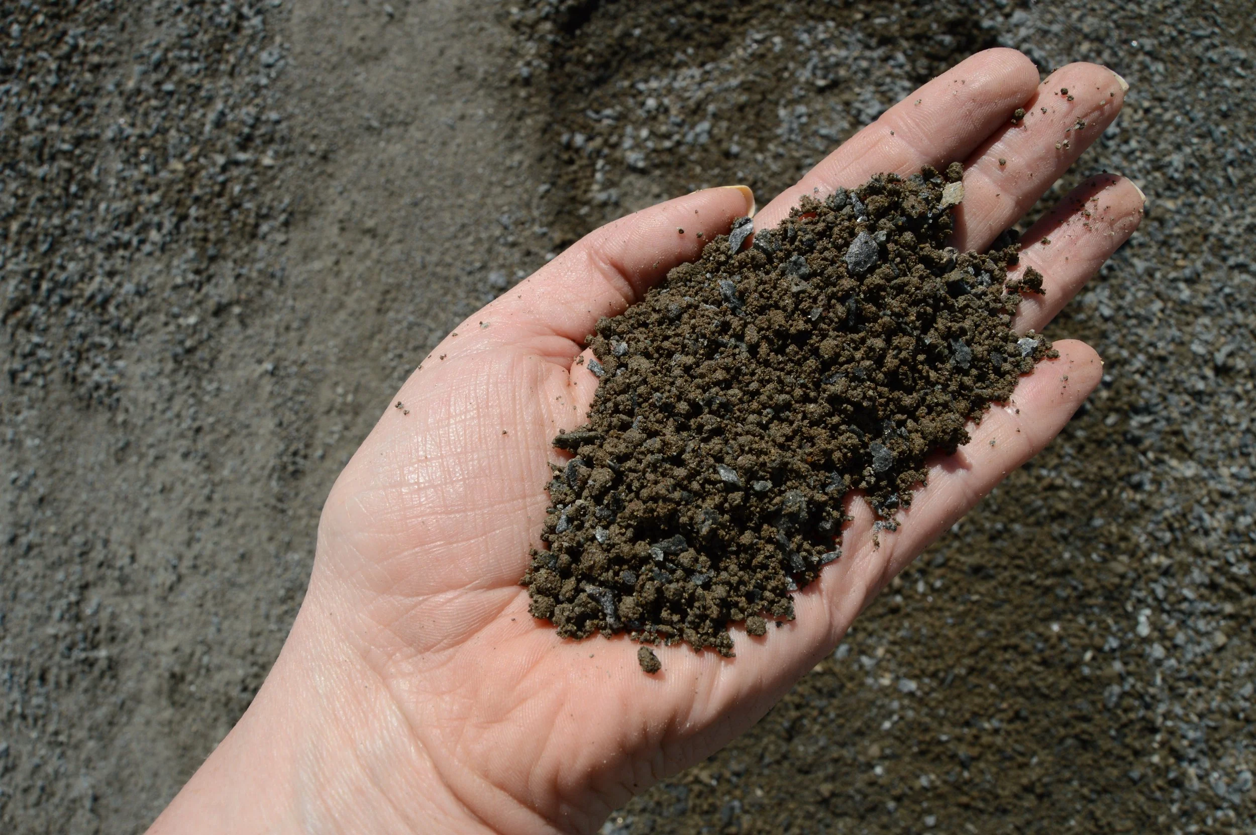 A hand holding a handful of dark, coarse soil or sand, with more sand on the ground.