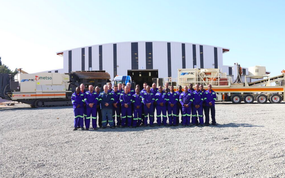Group of workers in blue uniforms standing in front of industrial equipment and a large building.