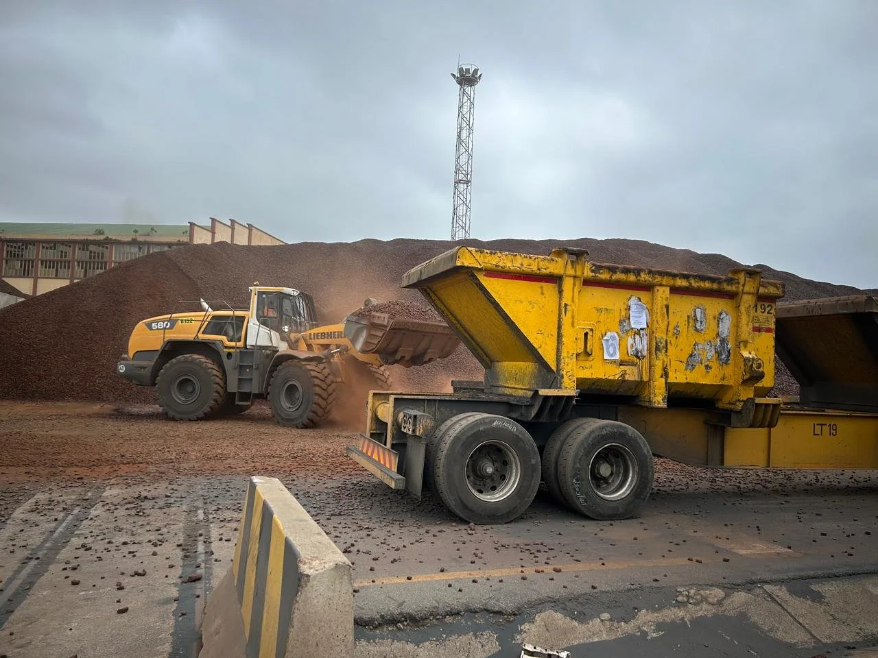 Heavy equipment working on a construction site, with a yellow dump truck and a large yellow front loader moving dirt or gravel, amid a cloudy sky and a landscape with a large pile of dirt in the background.