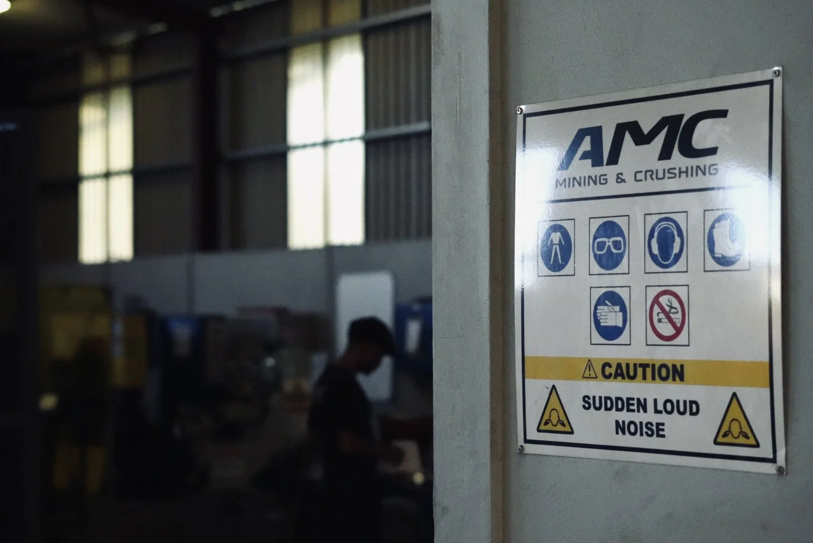 A safety sign for AMC Mining & Crushing on a wall, indicating safety precautions such as wearing a high-visibility vest, safety goggles, ear protection, and warning about sudden loud noise, as well as no smoking.