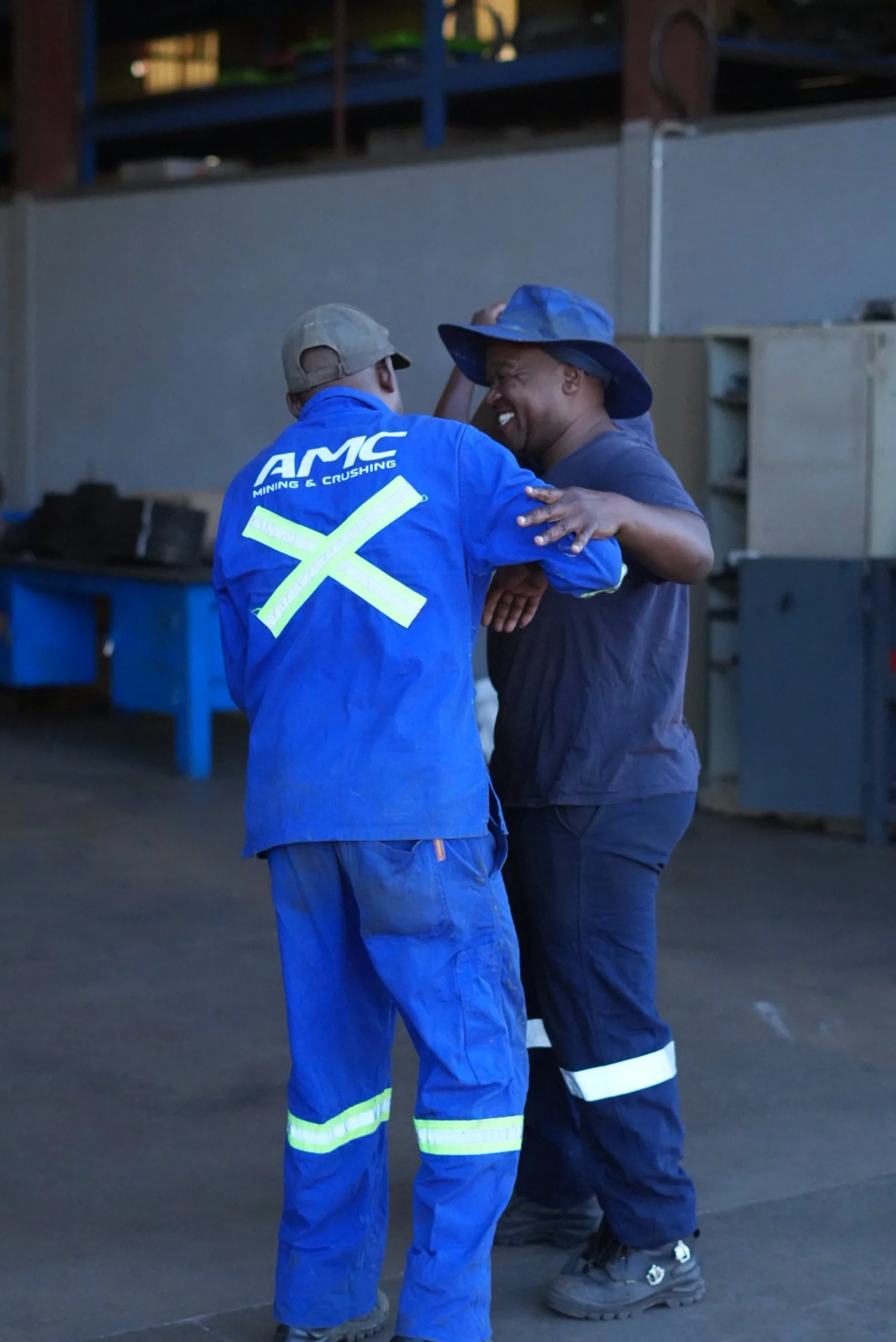 Two workers, one in blue coveralls with 'AMC Mining & Crushing' on the back, are smiling and embracing each other in a warehouse or industrial setting.