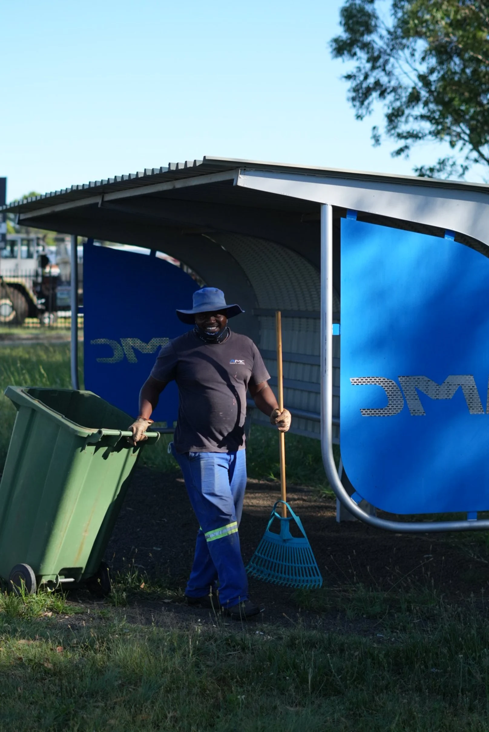 A person wearing blue work pants, a dark T-shirt, and a large blue hat is emptying a green trash bin outside, near a shelter, holding a broom and smiling at the camera.