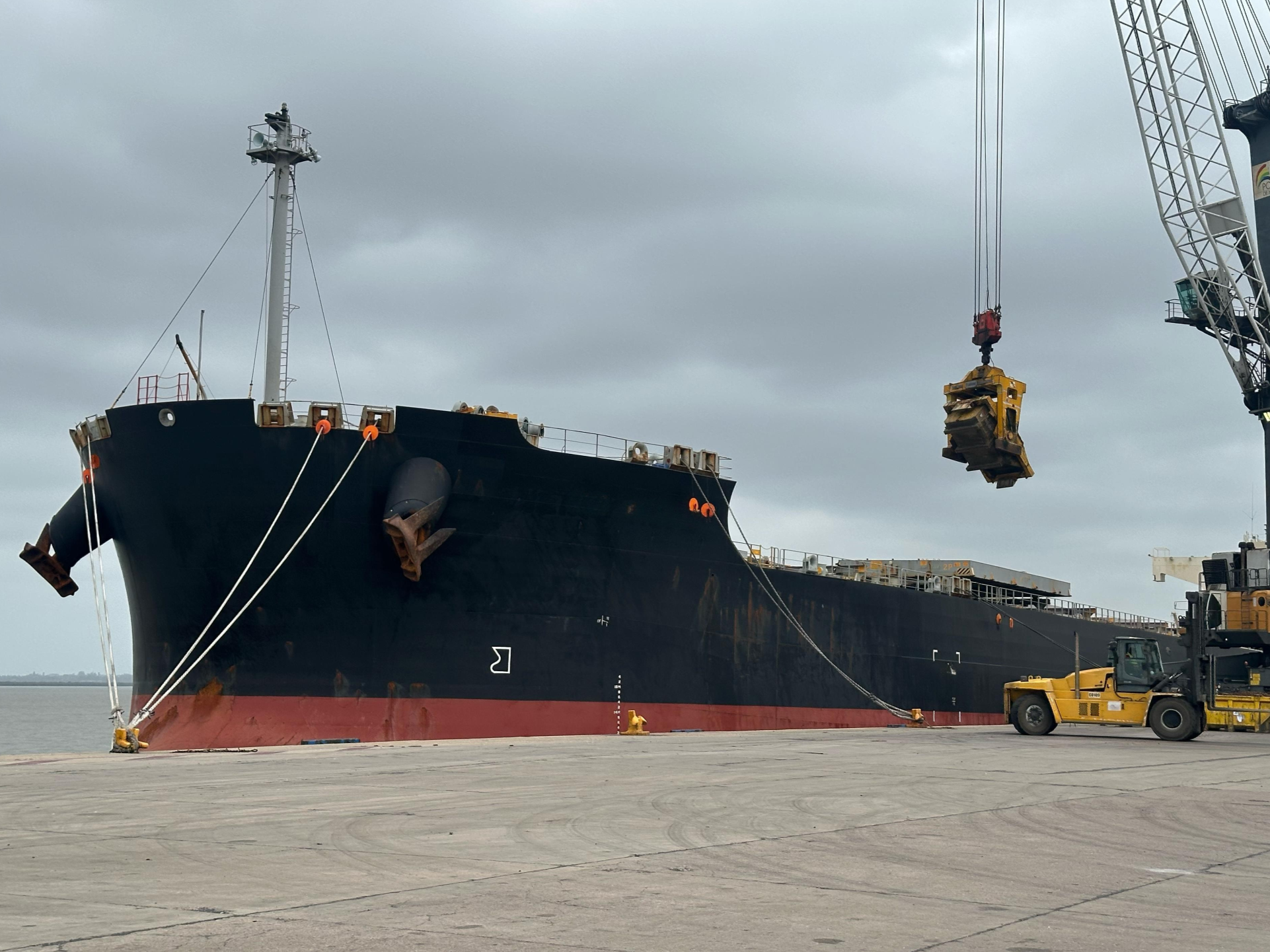 Large cargo ship docked at port with a crane lifting a yellow container, cloudy sky overhead.