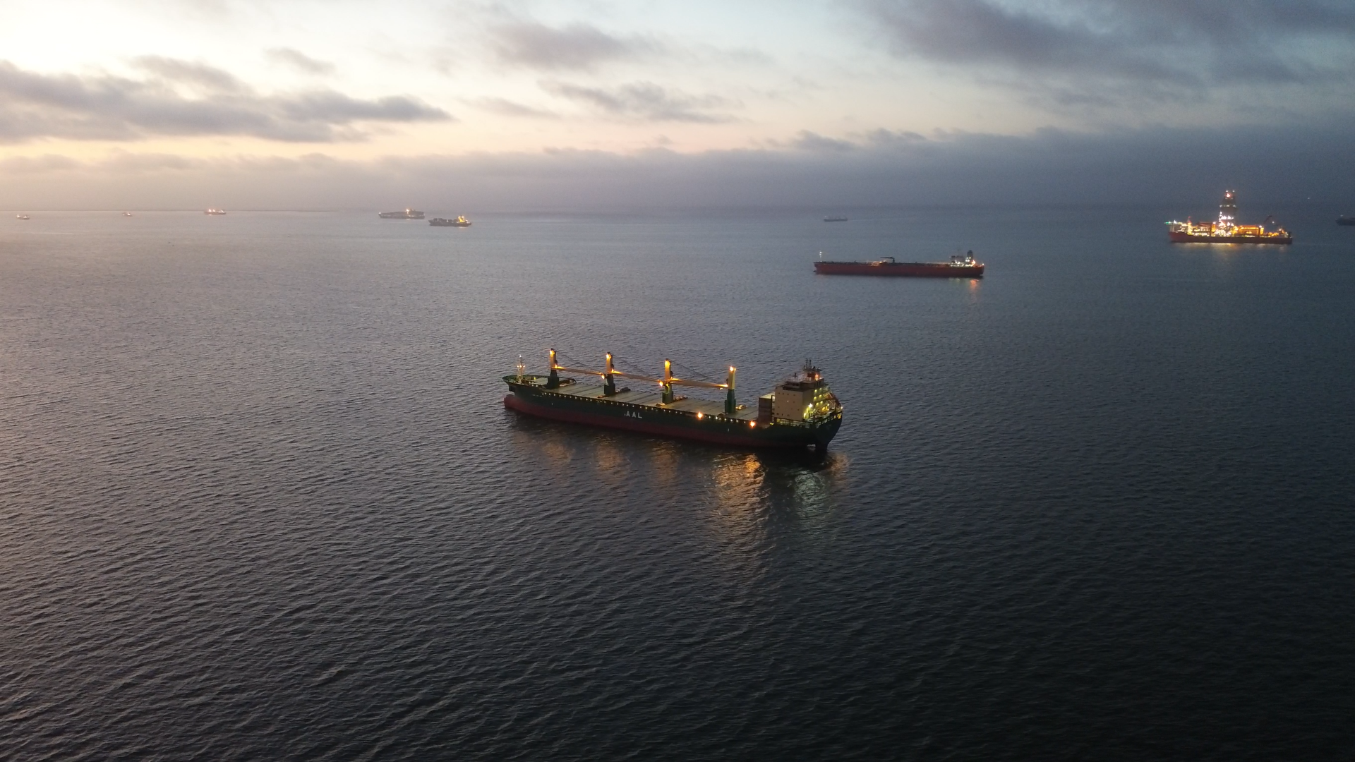 Several large ships floating on calm water during dusk, with a cloudy sky in the background.