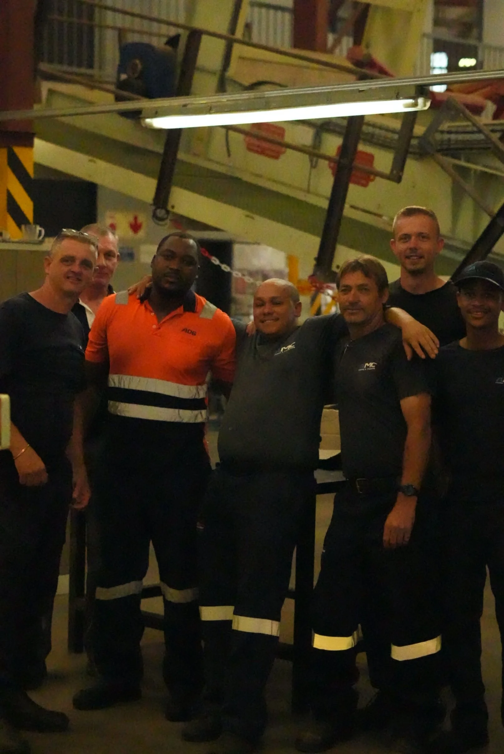 Group of seven people standing together inside an industrial or manufacturing facility, with some wearing uniforms and others casual clothing, posing for the photo.